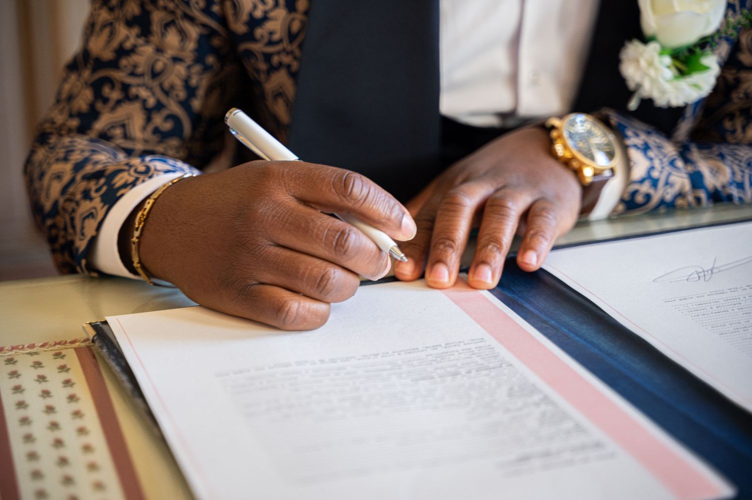 Close-up of groom signing wedding certificate with gold watch and floral suit jacket at ceremony.