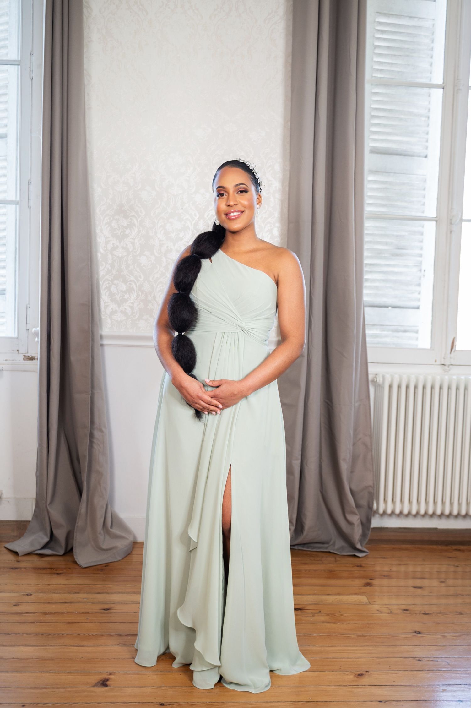 Elegant bridesmaid in mint green one-shoulder gown with long braided hair stands in a bright white room.