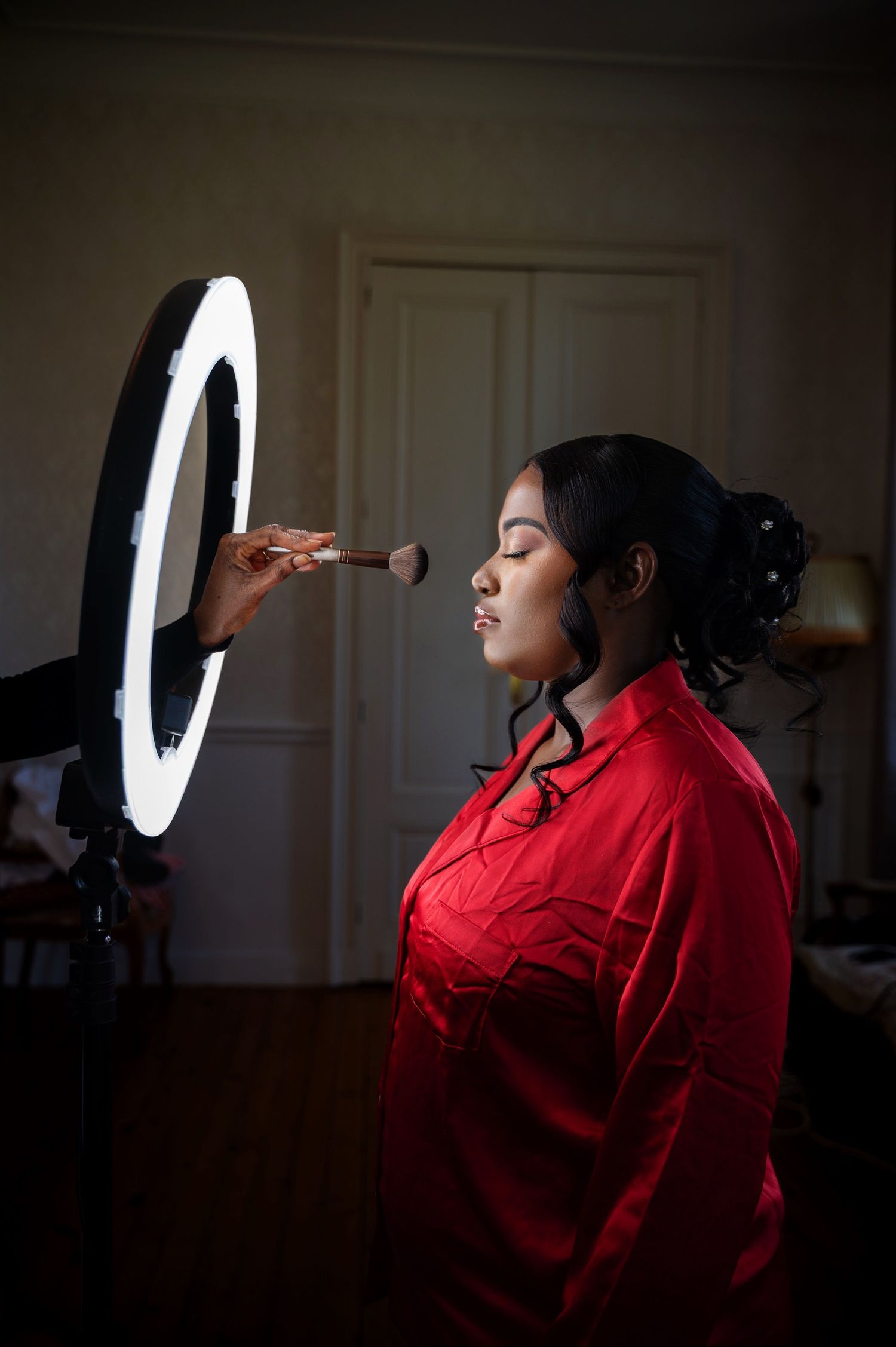 Bride in red satin robe getting makeup applied with brush under ring light before wedding ceremony.