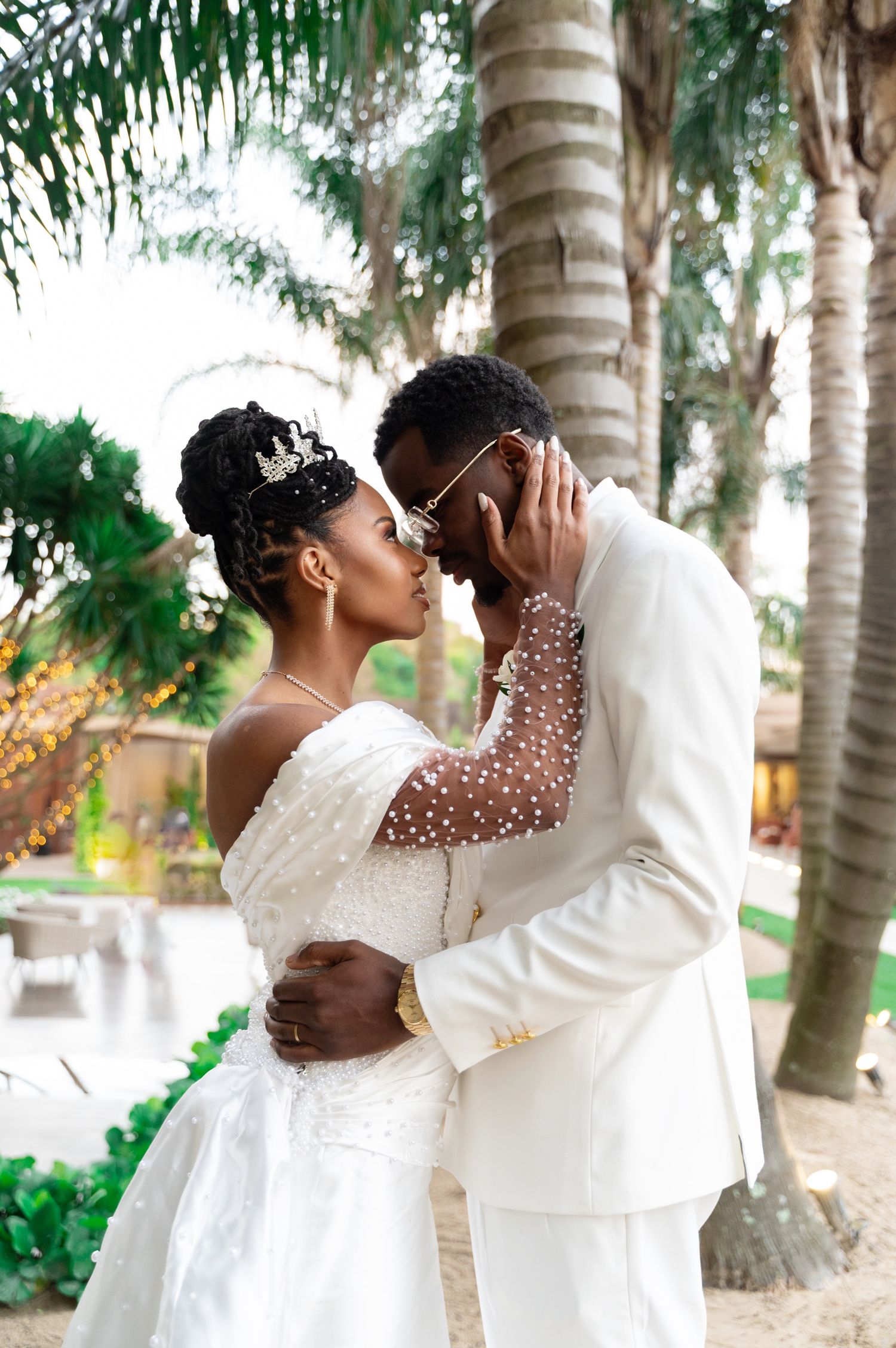 A Black bride and groom in white wedding attire share a romantic kiss under palm trees outdoors.