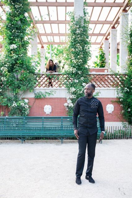 Man in black outfit stands in a lush garden with pergola, roses, and green benches in Paris.