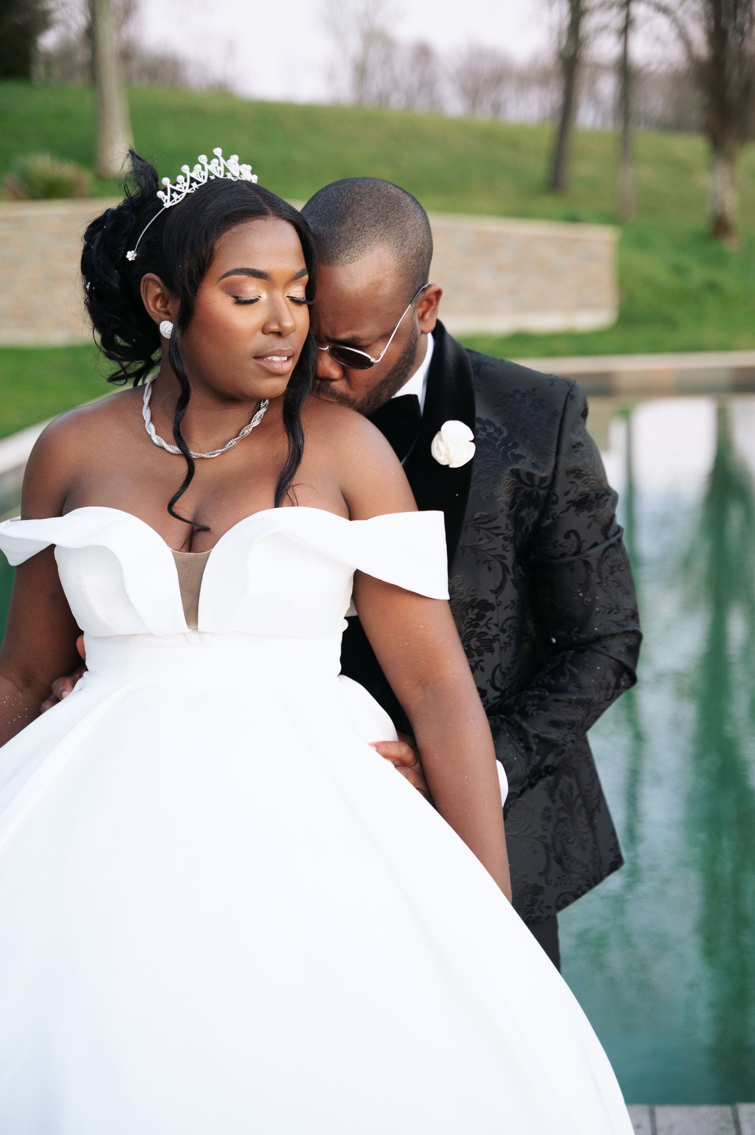 Groom in black tuxedo tenderly embraces bride in white ballgown near a reflective pool outdoors.