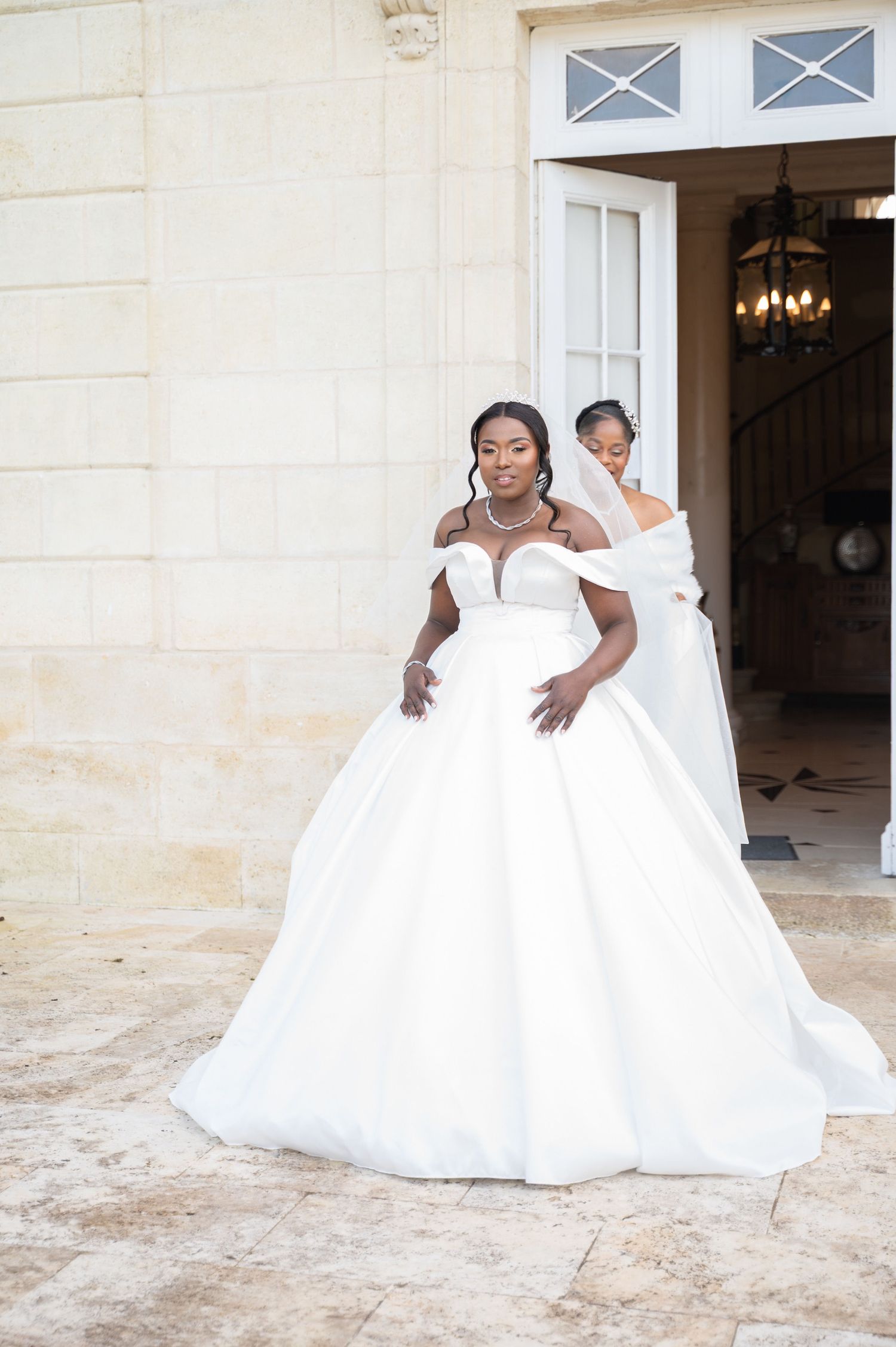 Black bride in stunning off-shoulder white ballgown stands outside a stone chateau with bridesmaid behind her.