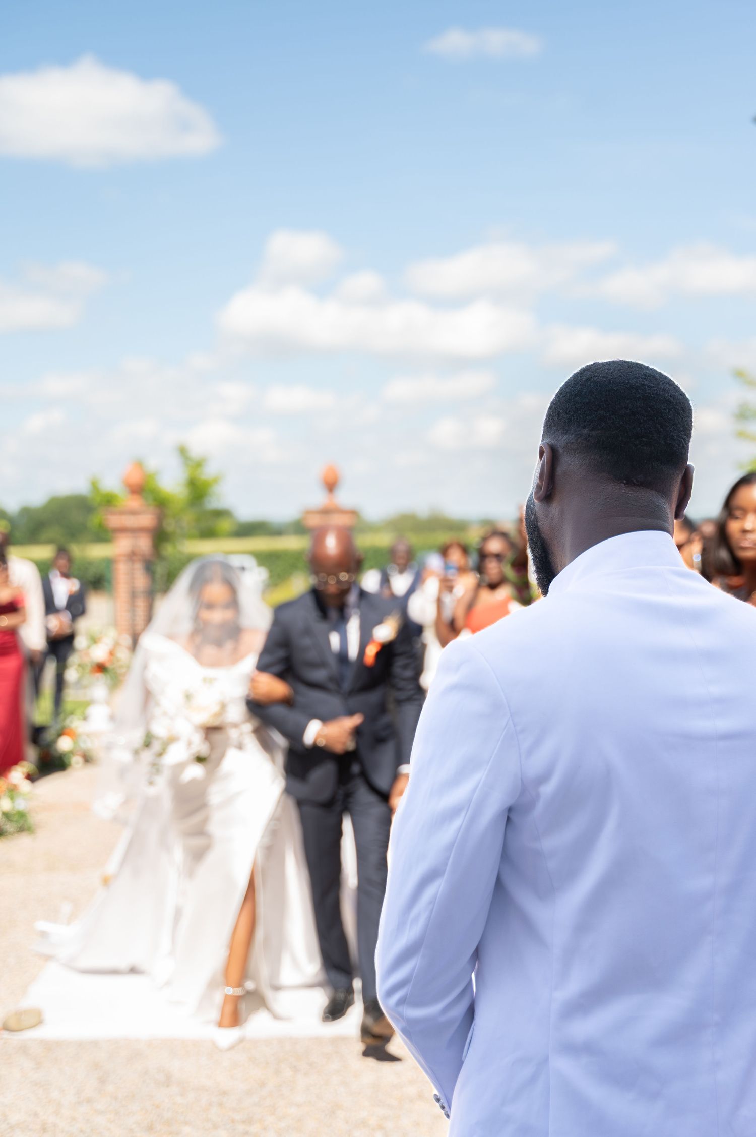 Groom in white suit watches bride approach down outdoor wedding aisle.