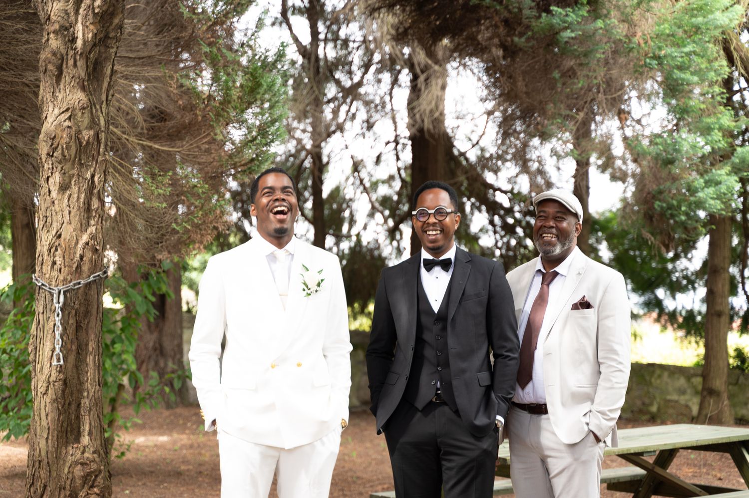Groom in white suit laughing with two groomsmen outdoors among tall pine trees.