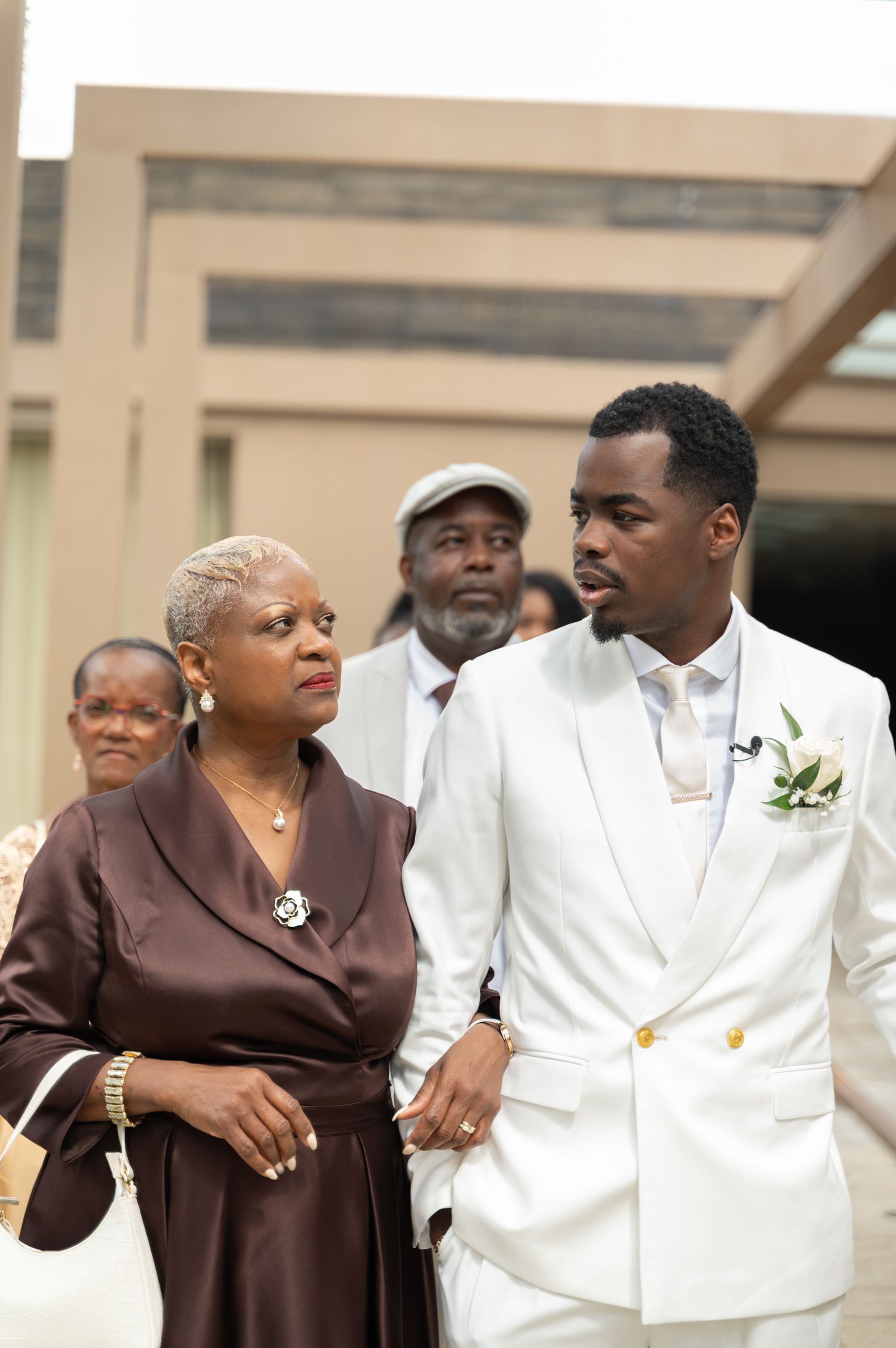 Groom in white suit with boutonniere walks with an older woman in brown satin at a wedding.