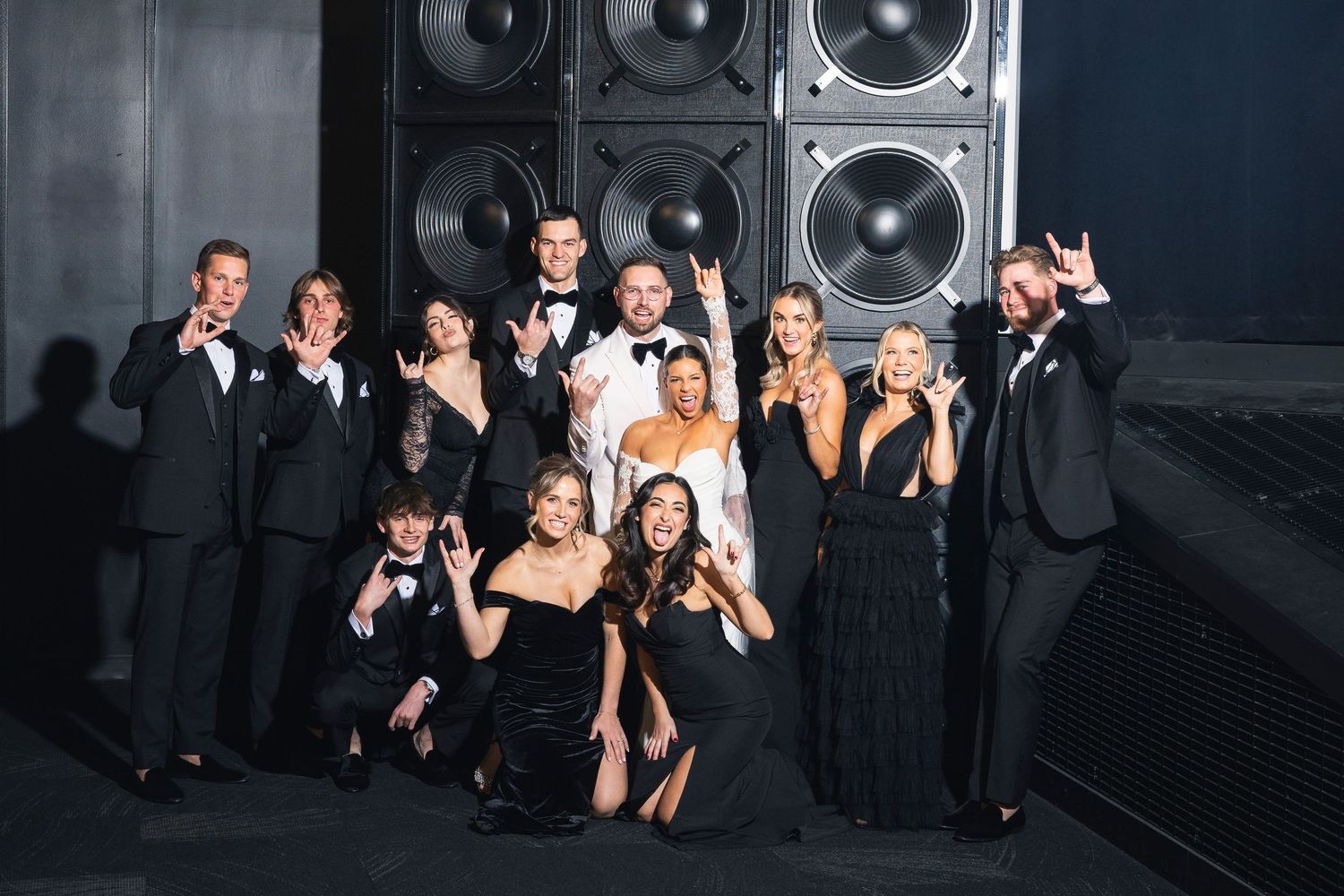 Wedding party posed together in formal attire against dark background with speakers.