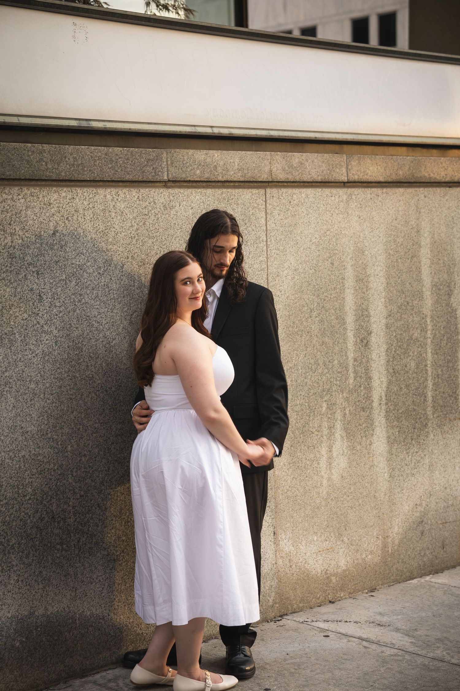 Two people in formal wear pose together against a textured stone wall during a photoshoot.