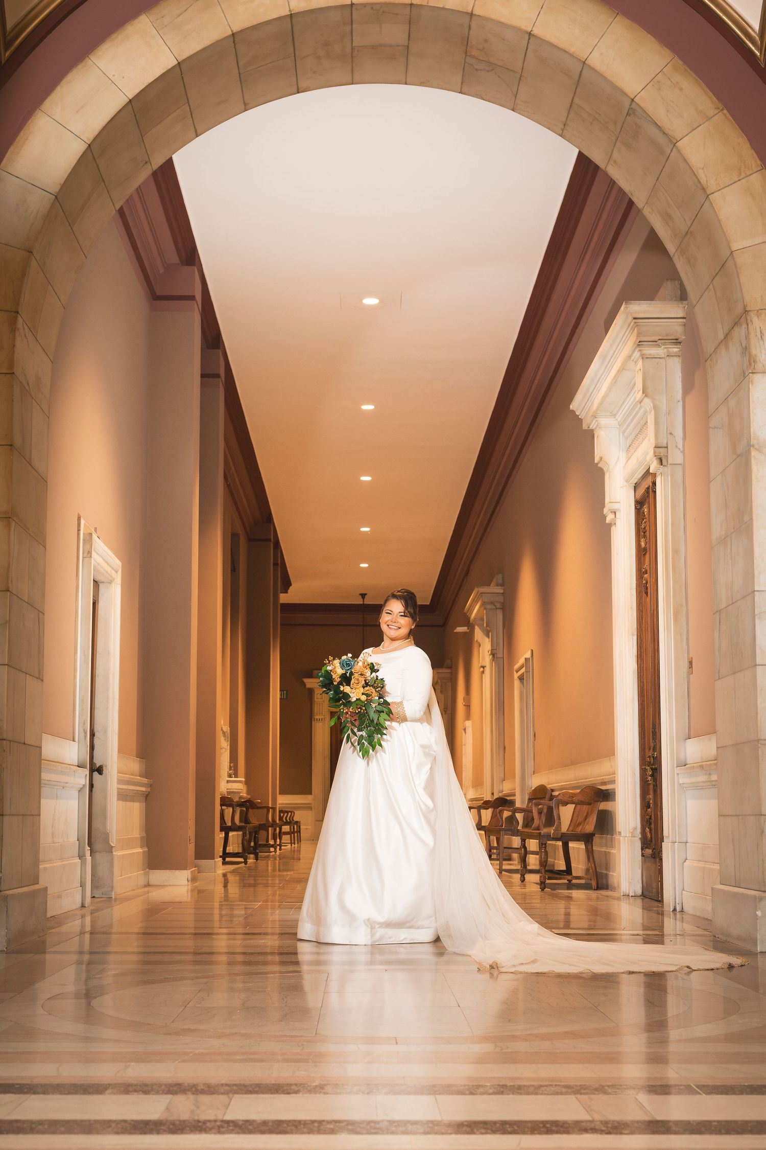 A bride in white dress poses in a grand corridor with arched stone architecture.