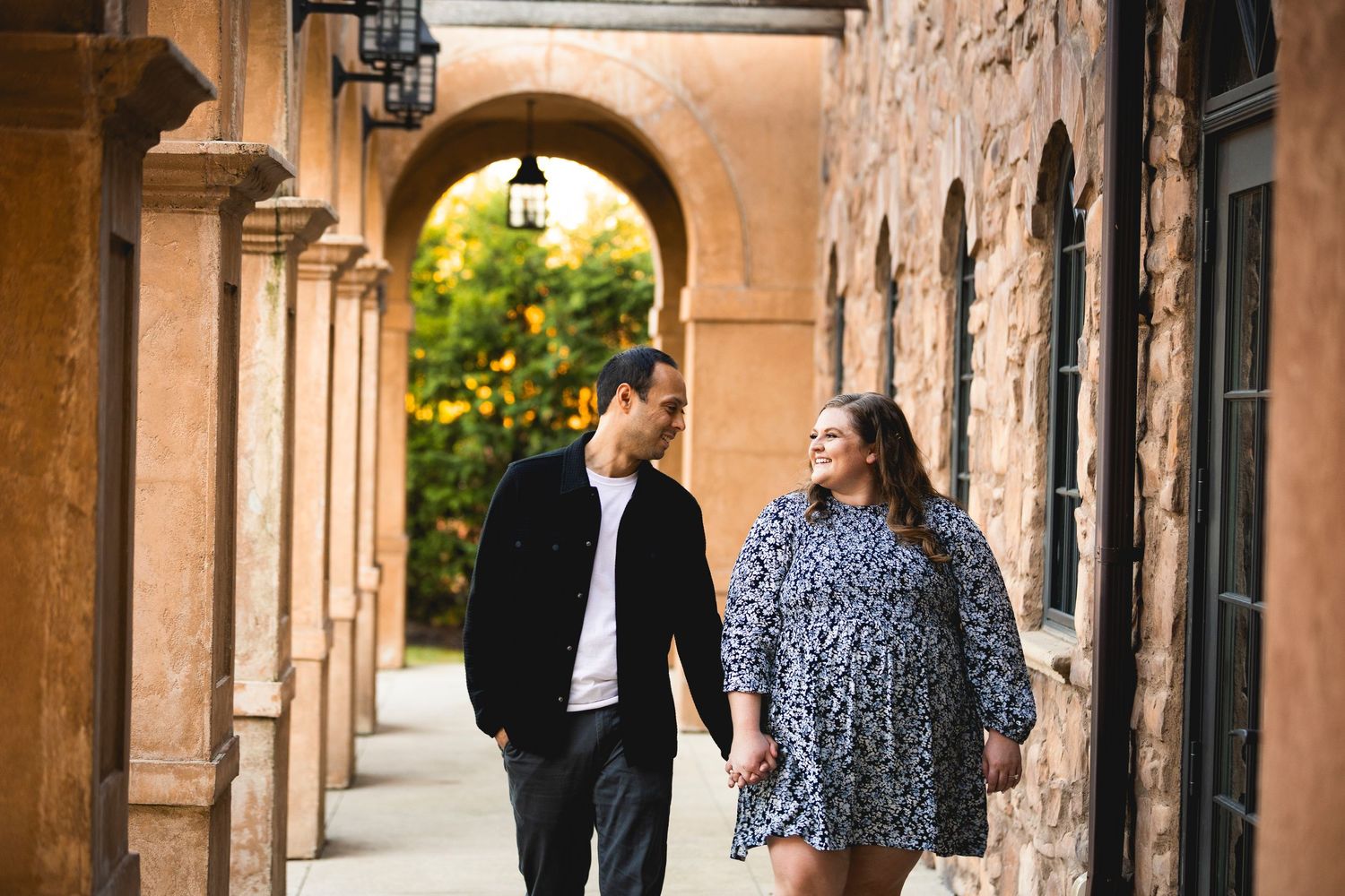 A couple strolls through an elegant stone archway corridor with vintage architecture and warm lighting.