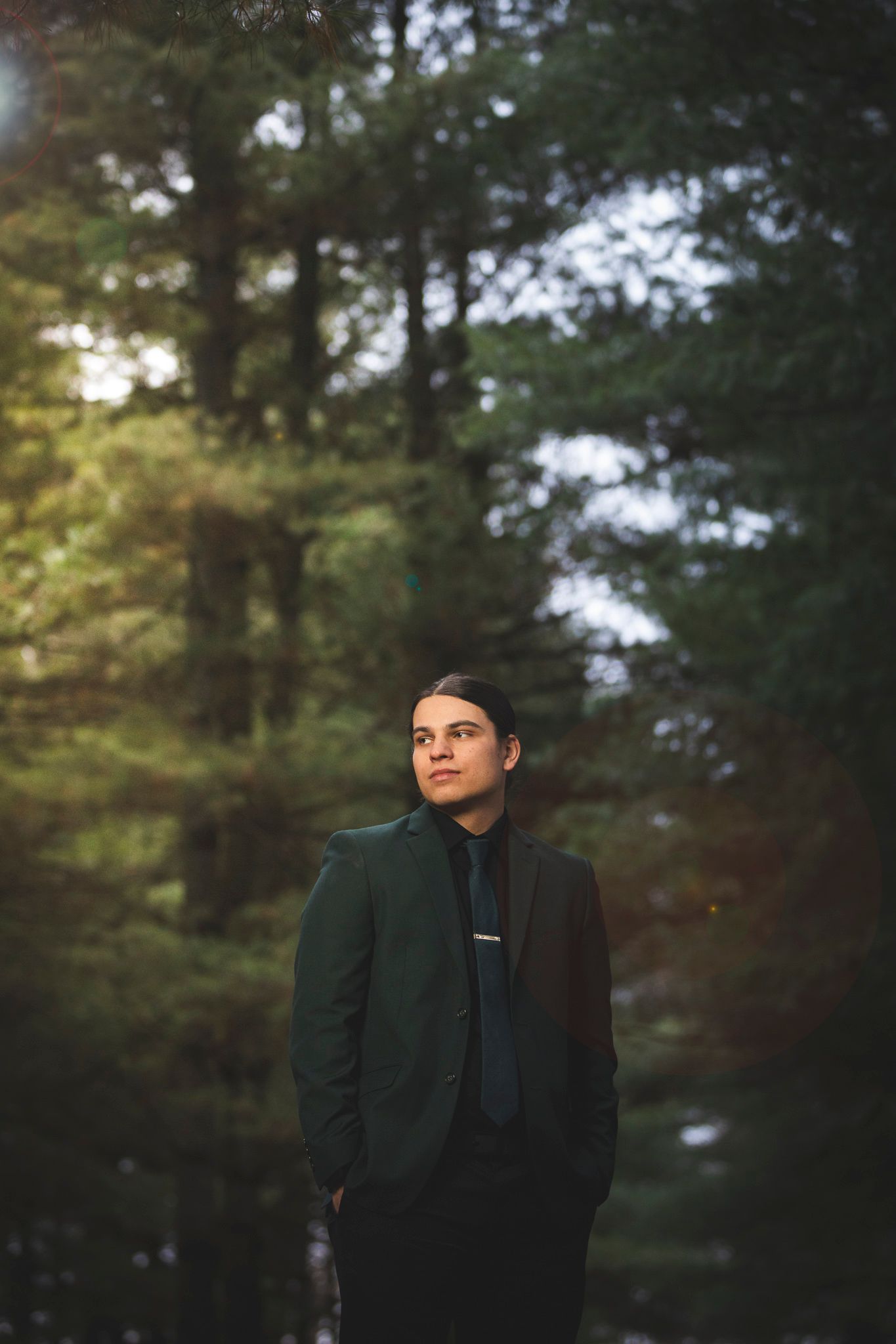 A person in a dark green formal jacket poses thoughtfully in front of a wooded outdoor setting at dusk.