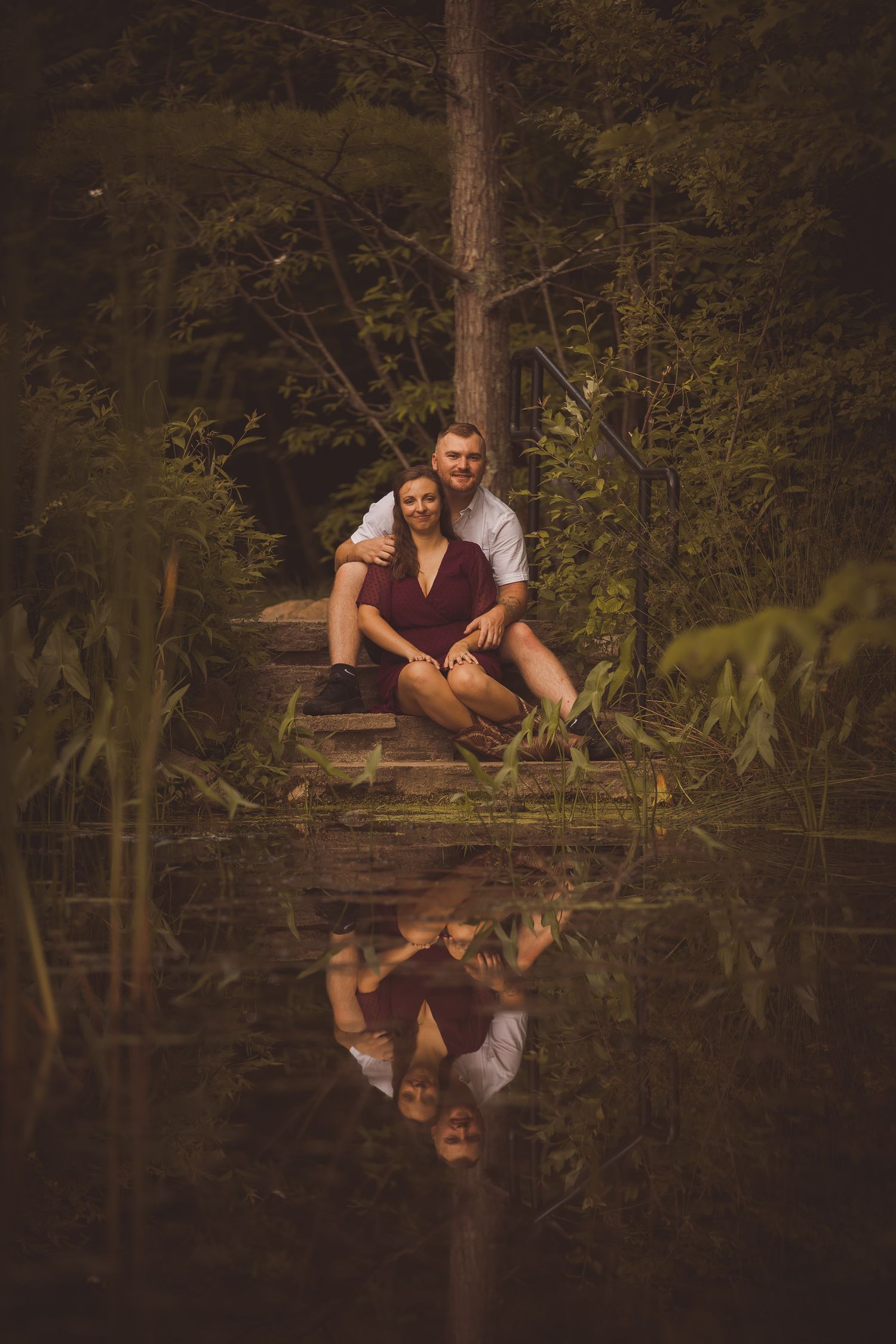 A couple sits on a wooden dock reflecting in still water surrounded by trees.