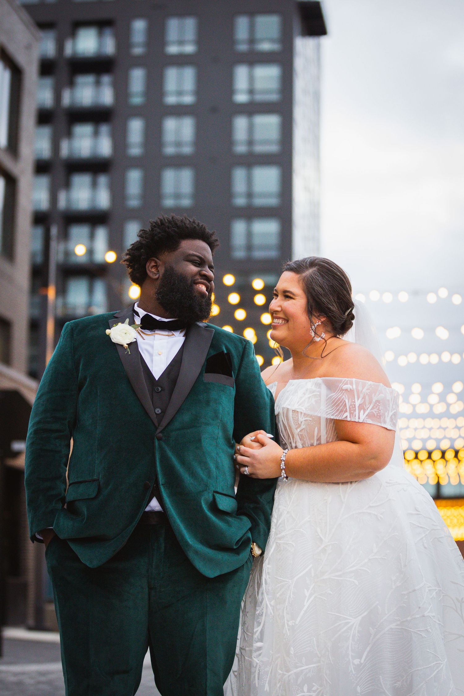 Wedding couple shares joyful moment under twinkling lights in urban setting.