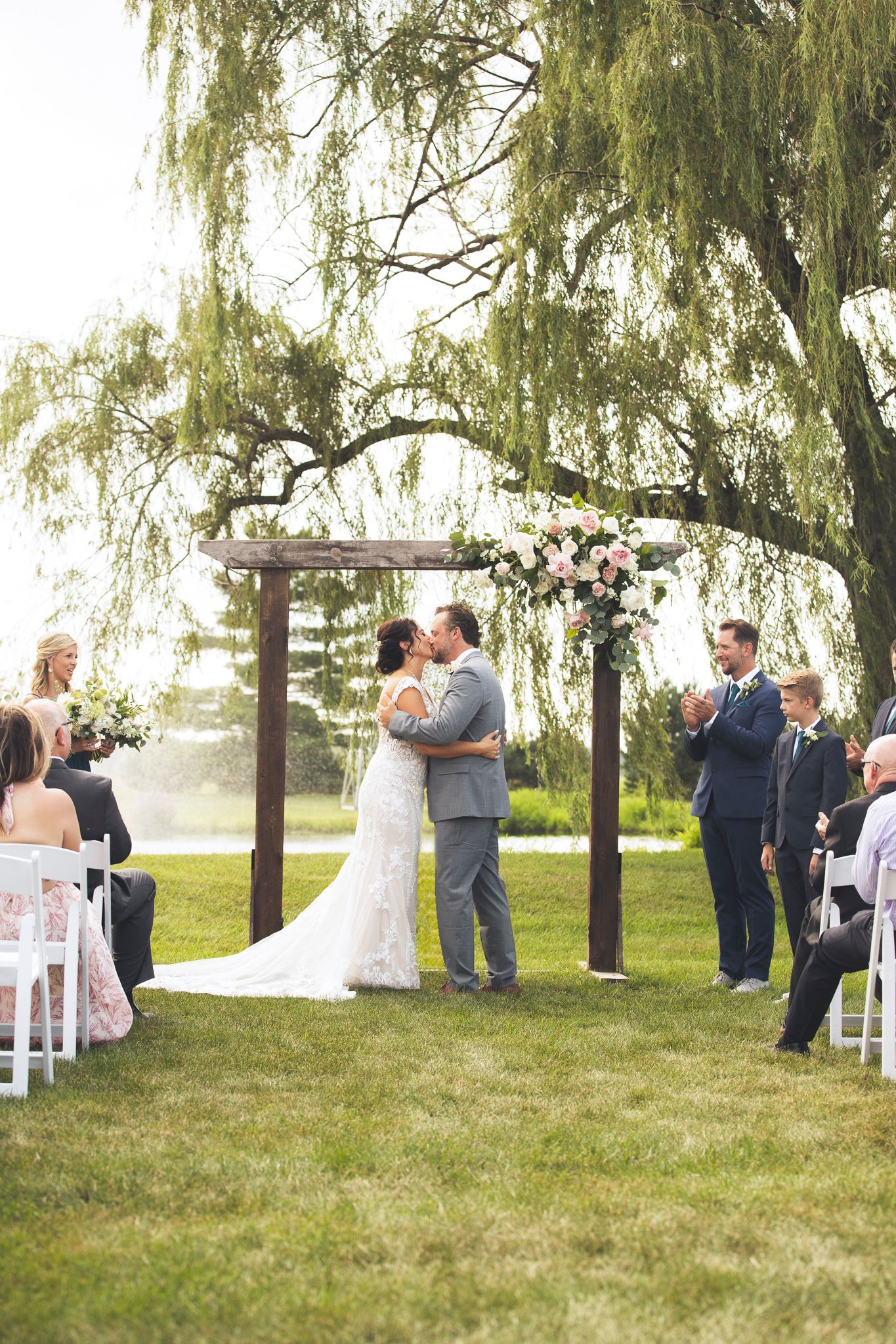 An outdoor wedding ceremony under weeping willow trees with guests seated on white chairs.