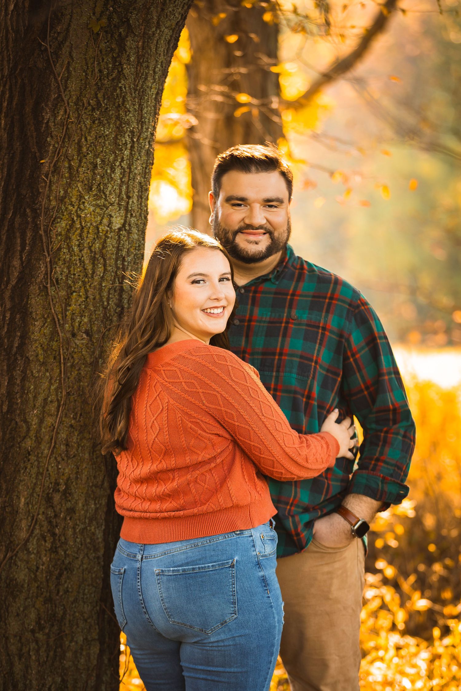 A couple poses playfully by a tree trunk in autumn with golden sunlight filtering through.