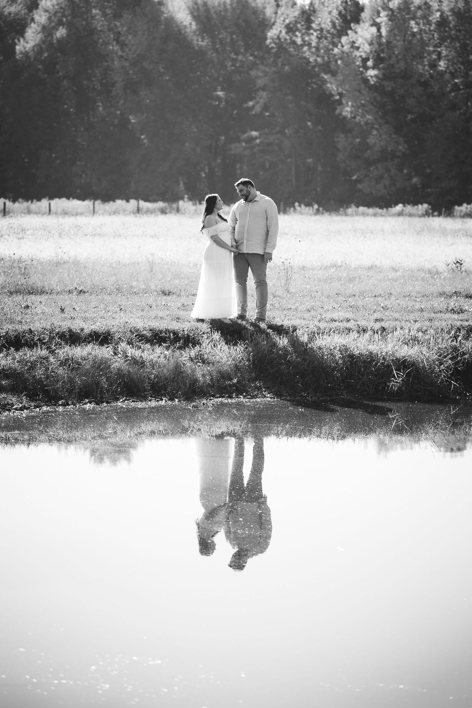 A black and white photograph captures two people's reflection in still water during sunset.