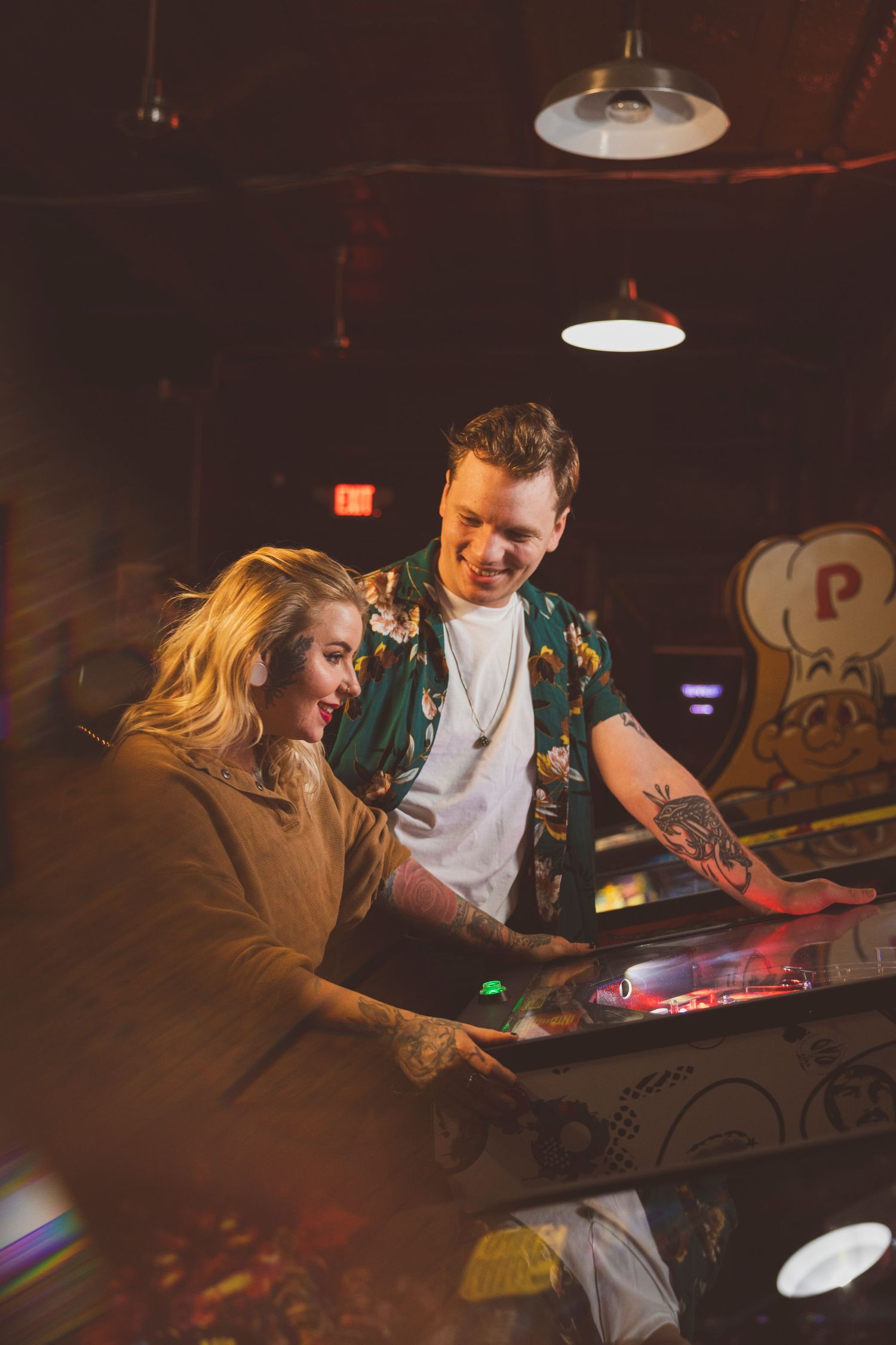 Couple leaning close together at bar with industrial lighting overhead.