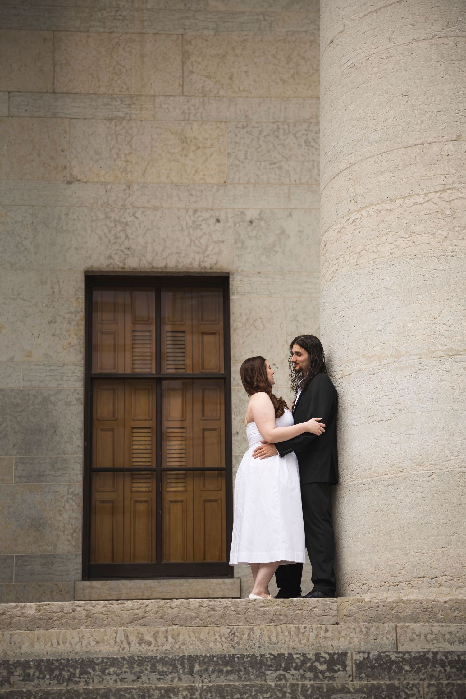 Wedding couple poses against textured stone wall with wooden windows in outdoor setting.