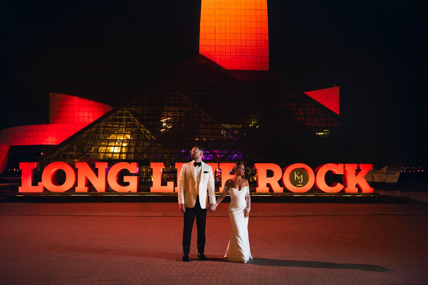 A couple stands silhouetted against a red neon sign reading 'LONG LIVE ROCK' at night.