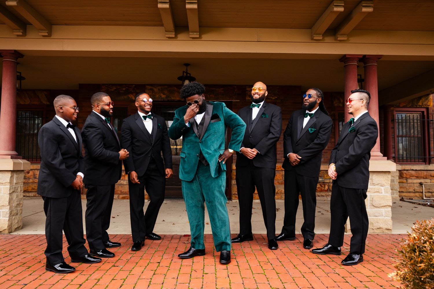 Groomsmen in emerald green and black suits pose in line outside brick building.