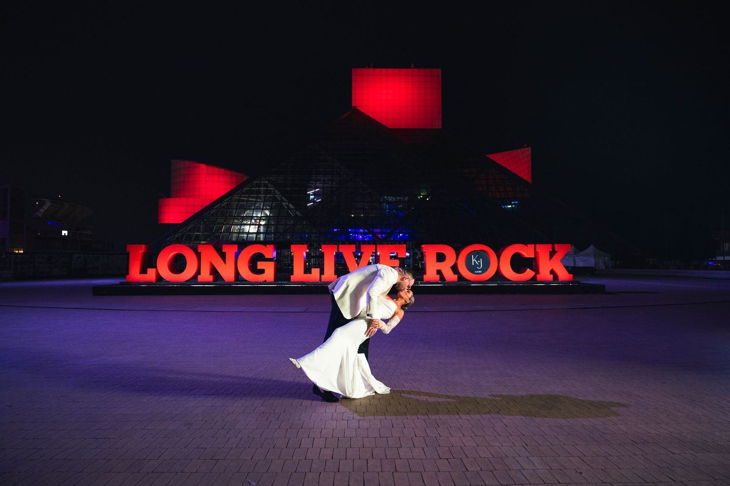 Person in white dress twirls in front of red-lit 'LONG LIVE ROCK' sign at night.