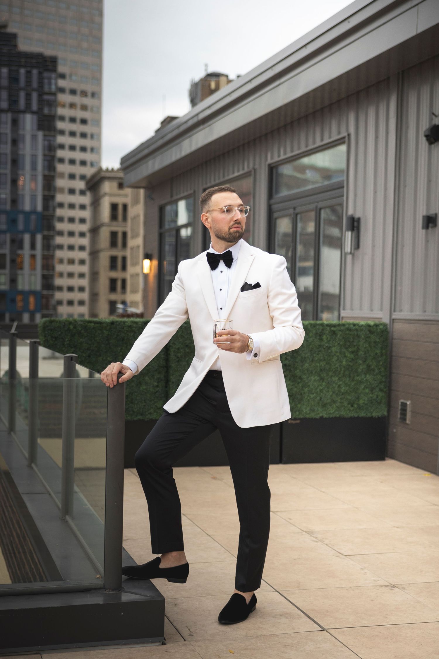 Person in white dinner jacket and black bow tie poses on modern rooftop terrace.