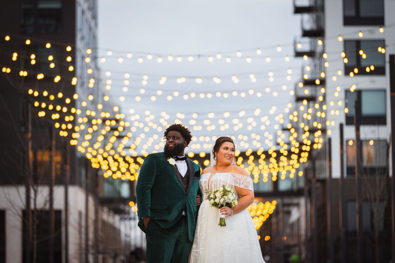 A romantic moment captured under glowing string lights in an urban setting at dusk.