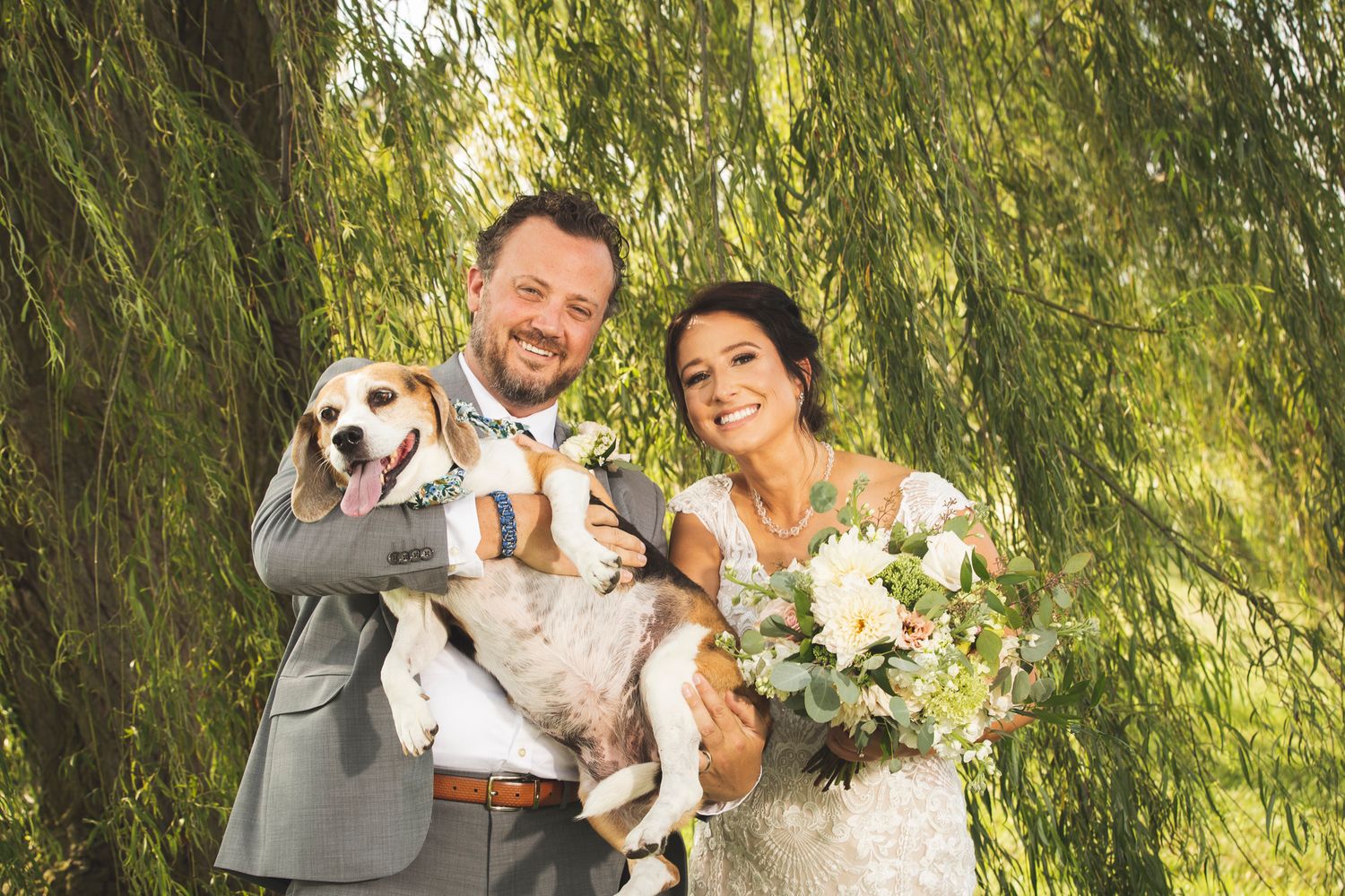A joyful wedding couple poses with their dog under a weeping willow tree in a park setting.