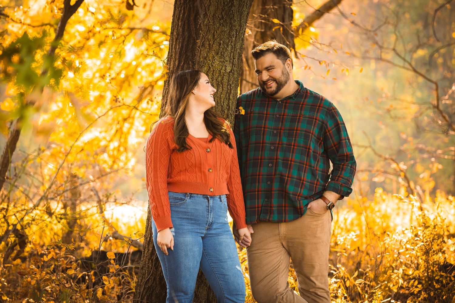 A couple shares laughter while leaning against a tree during a golden autumn afternoon.