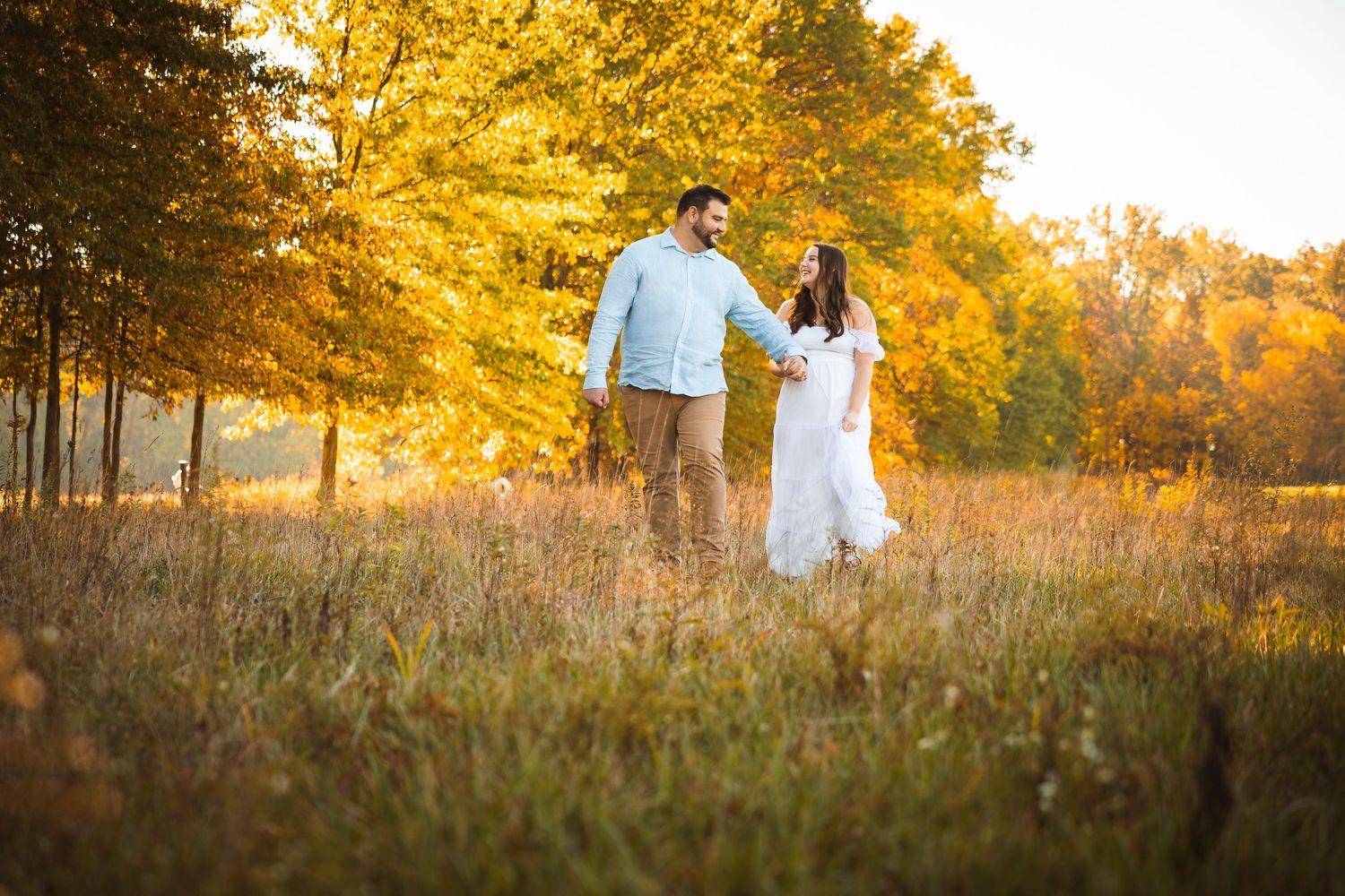Two people walk through a golden meadow with autumn trees creating a warm backdrop.