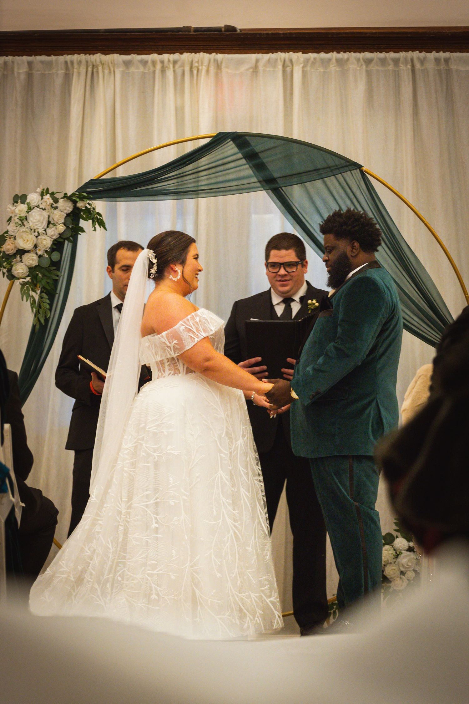 Intimate wedding ceremony moment under an emerald green and gold arch with white floral decorations.