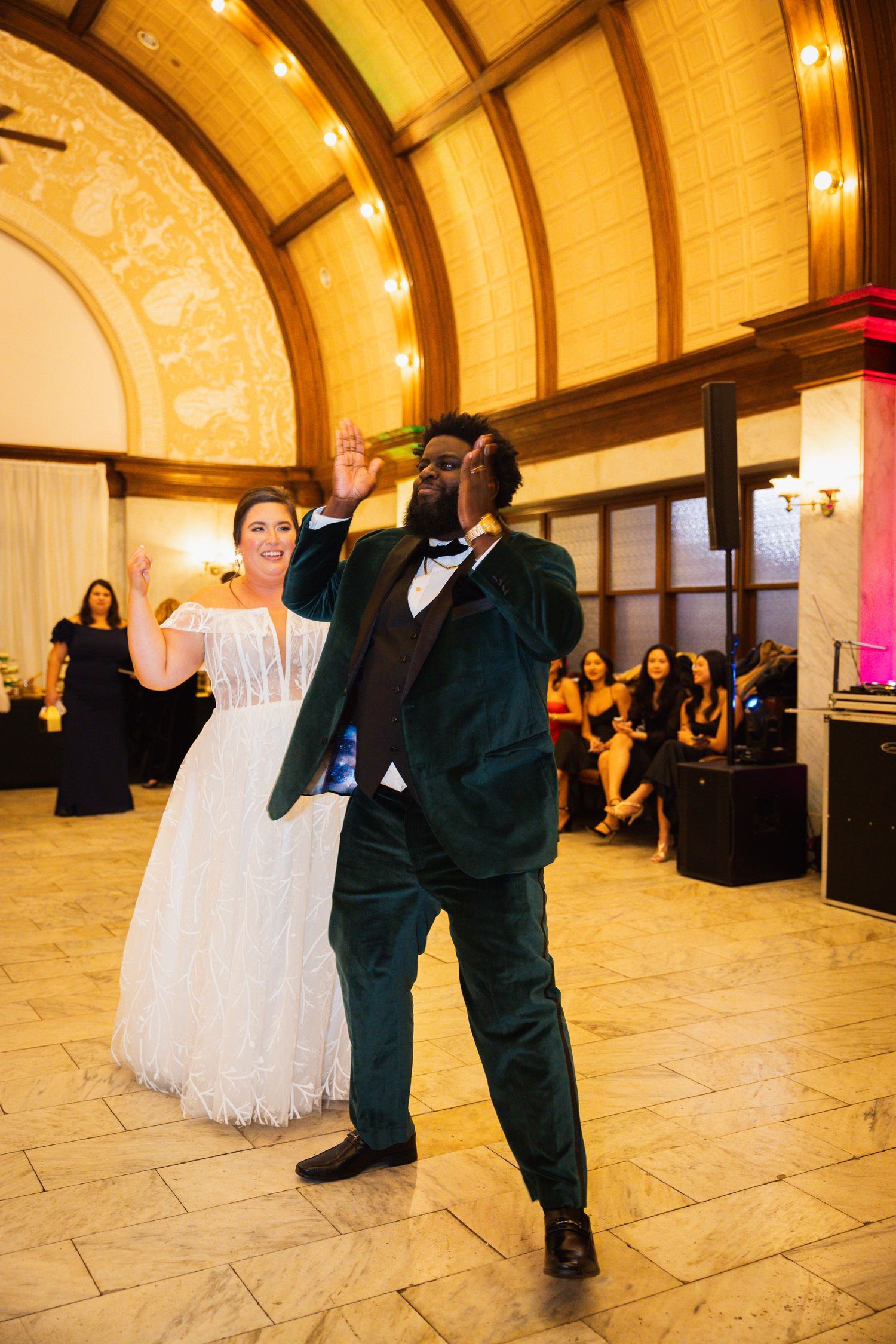A couple shares a dance at their wedding reception in an elegant ballroom with a vaulted ceiling and warm lighting.