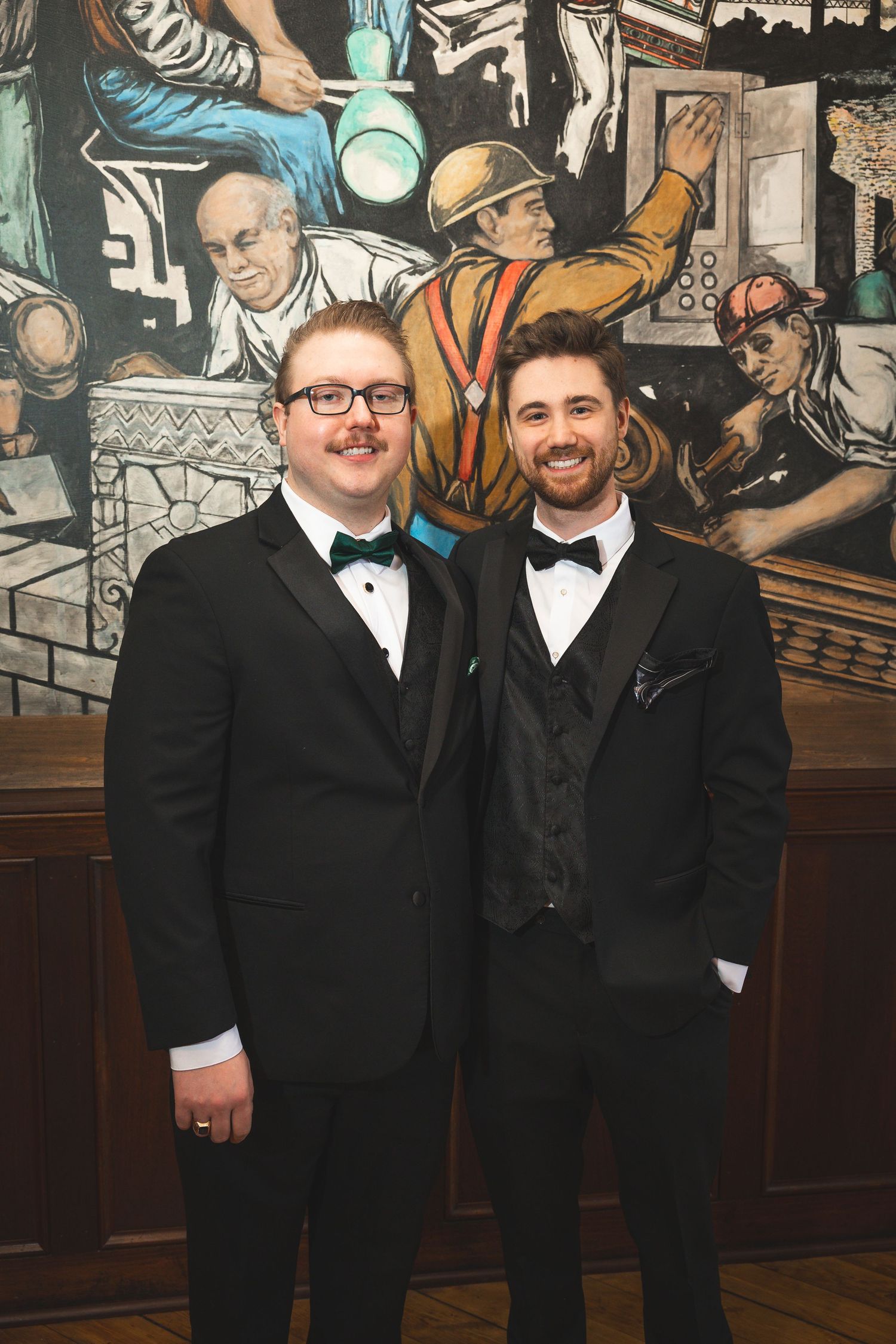 Wedding party members in formal black suits pose against decorative mural backdrop.