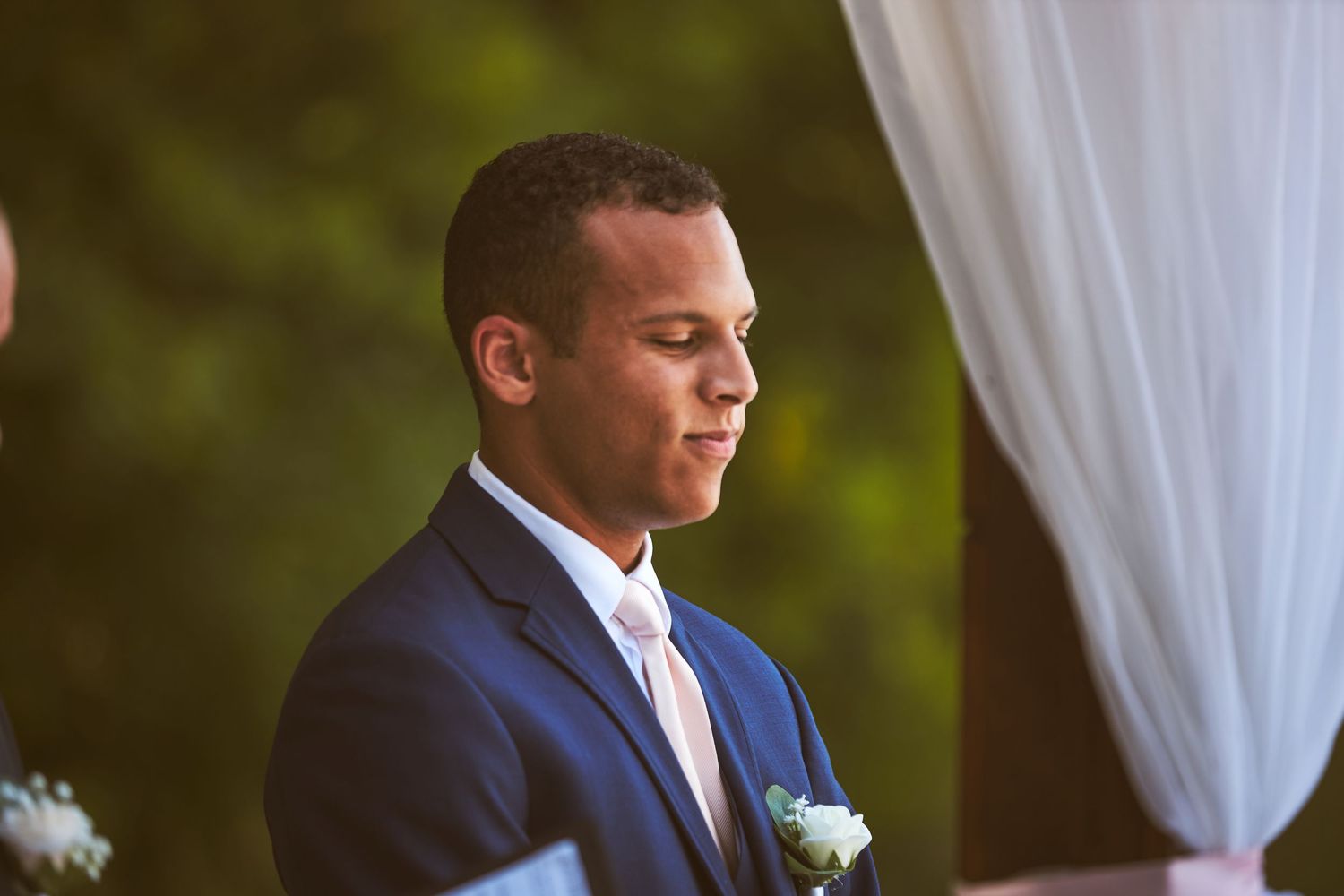 Groomsmen in navy suits listen attentively during an outdoor ceremony.