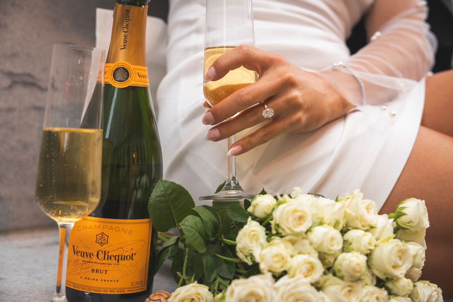 Champagne and white roses arranged for celebration against stone wall.