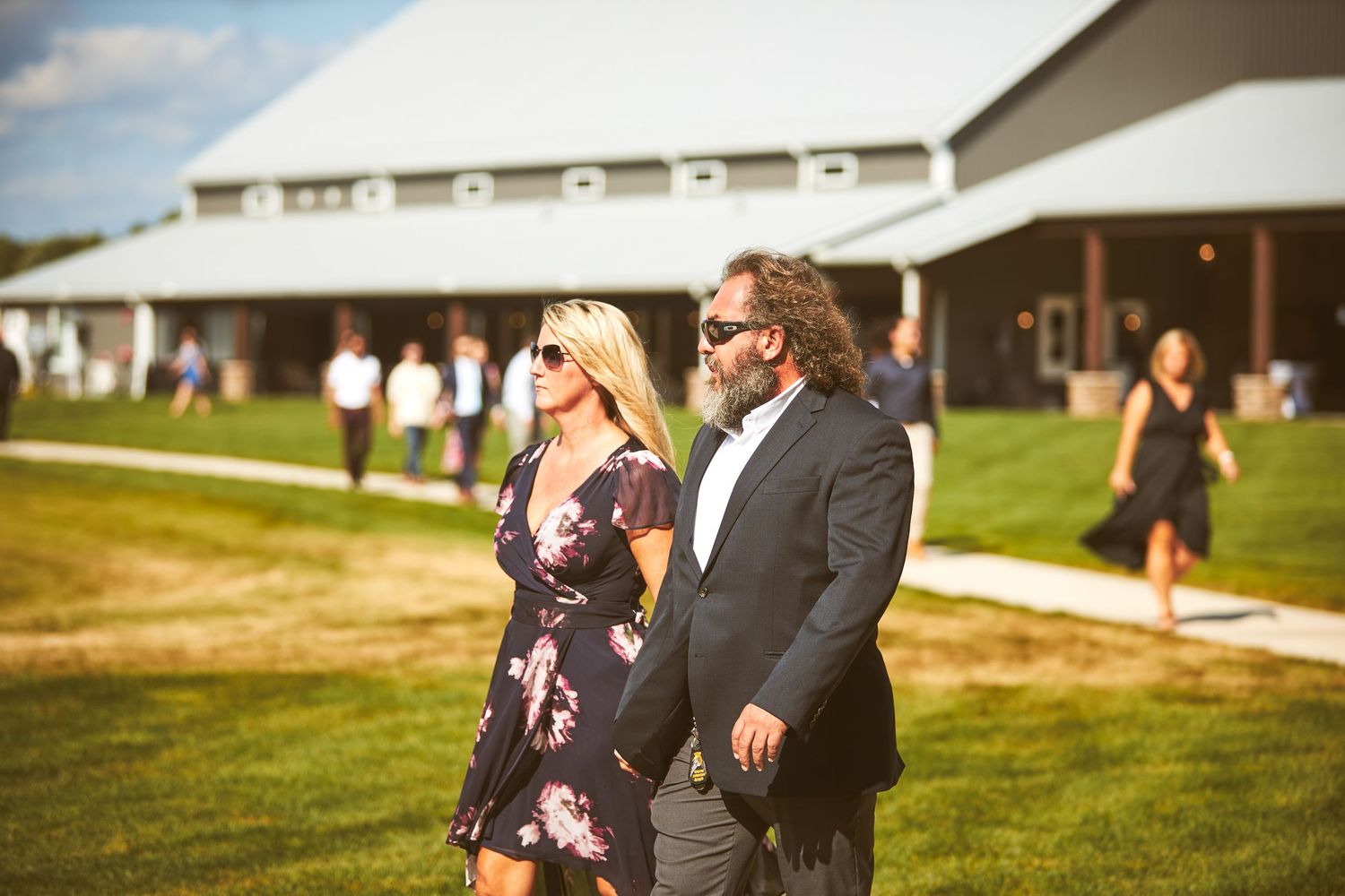 Wedding guests mingle outdoors on a sunny day near a building.