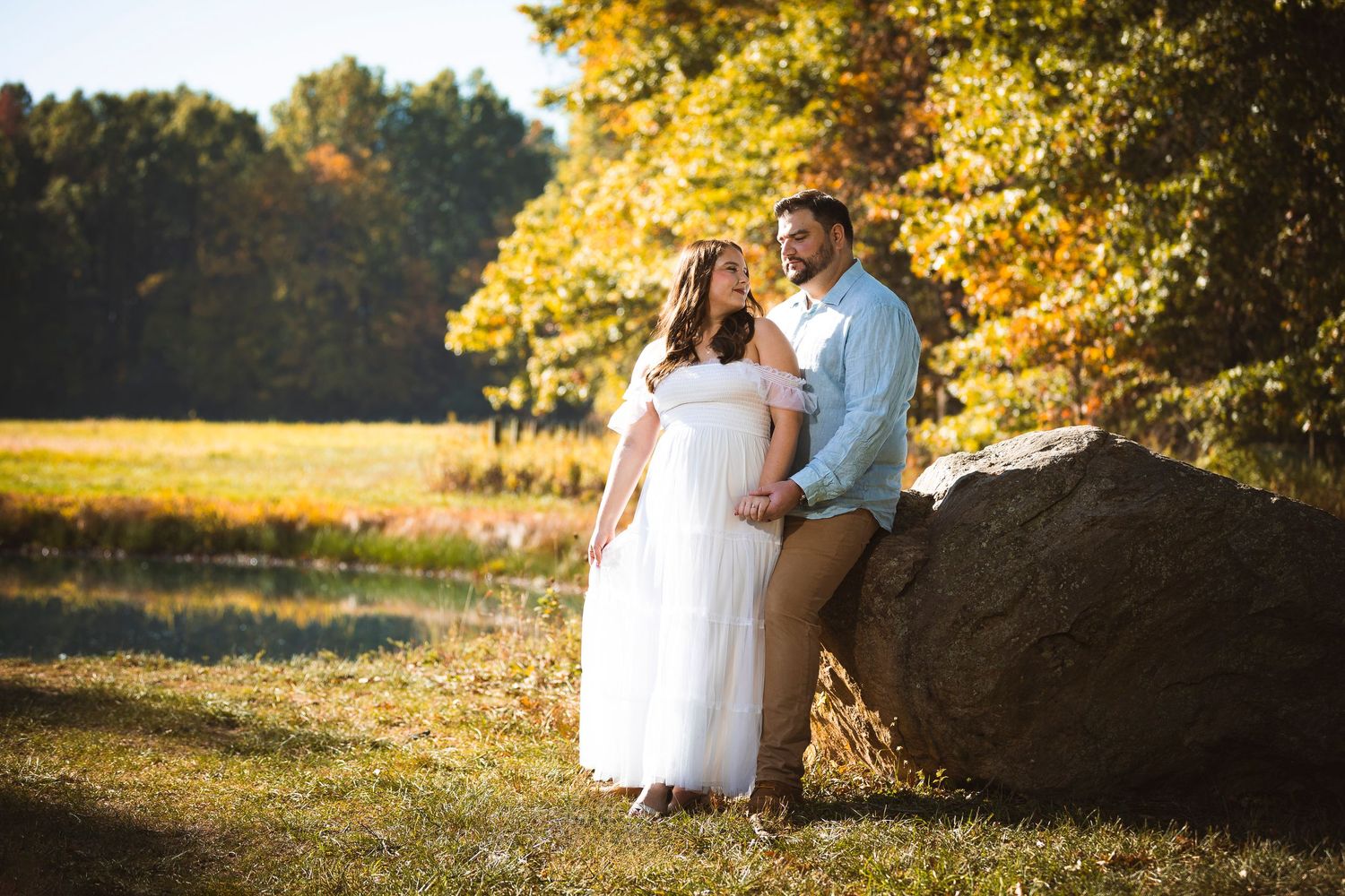 A couple sits together on large rocks surrounded by autumn foliage during golden hour.