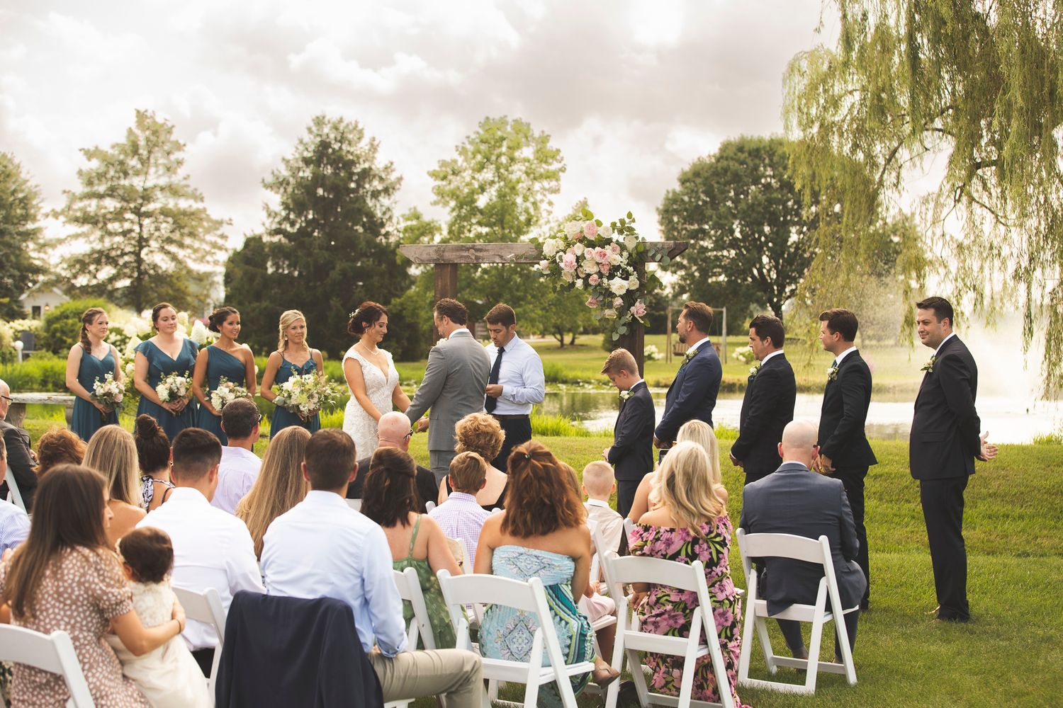 A wedding ceremony scene with guests seated outdoors under willow trees on a sunny day.