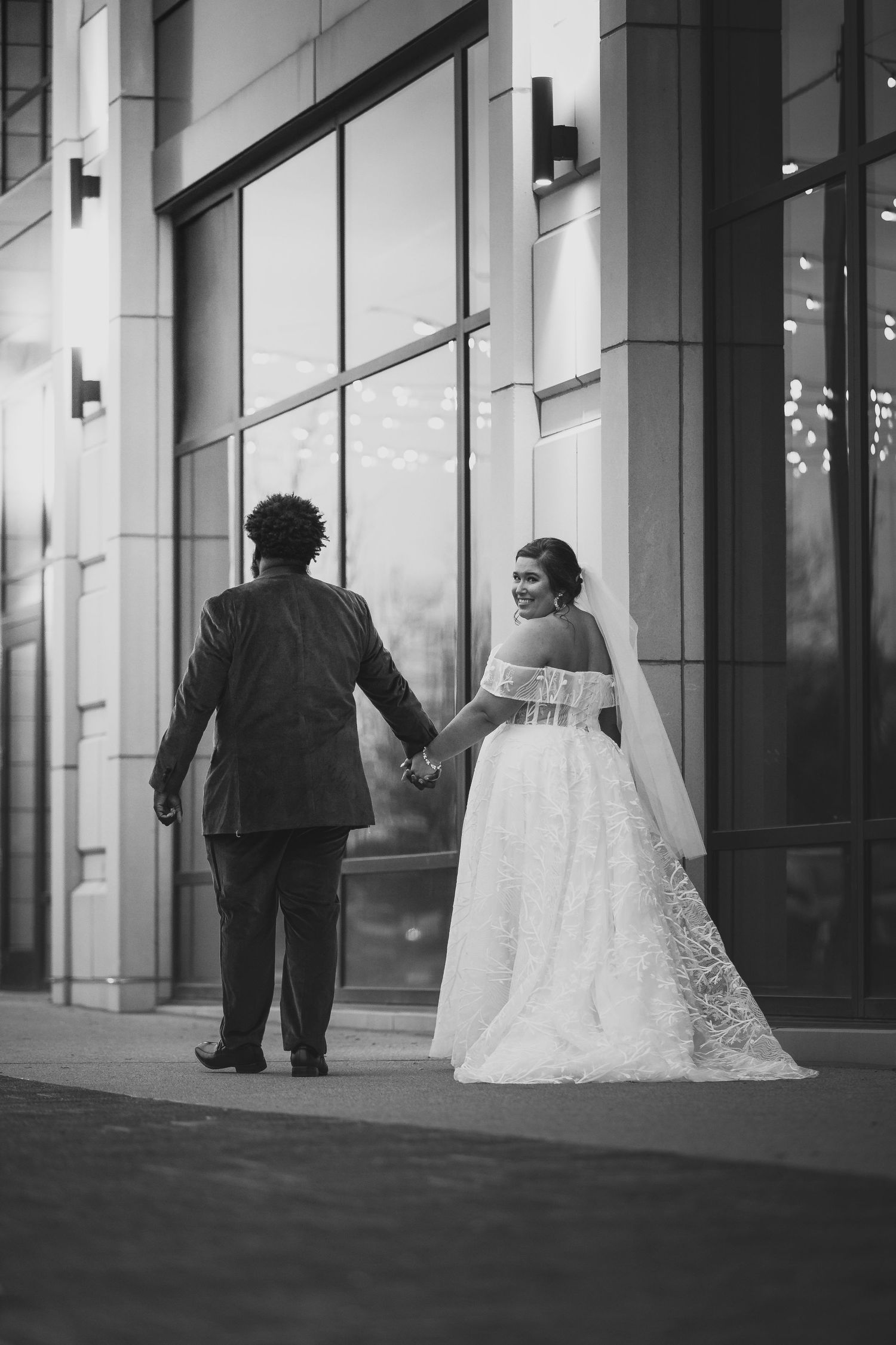 Black and white wedding portrait with couple walking along modern building exterior.