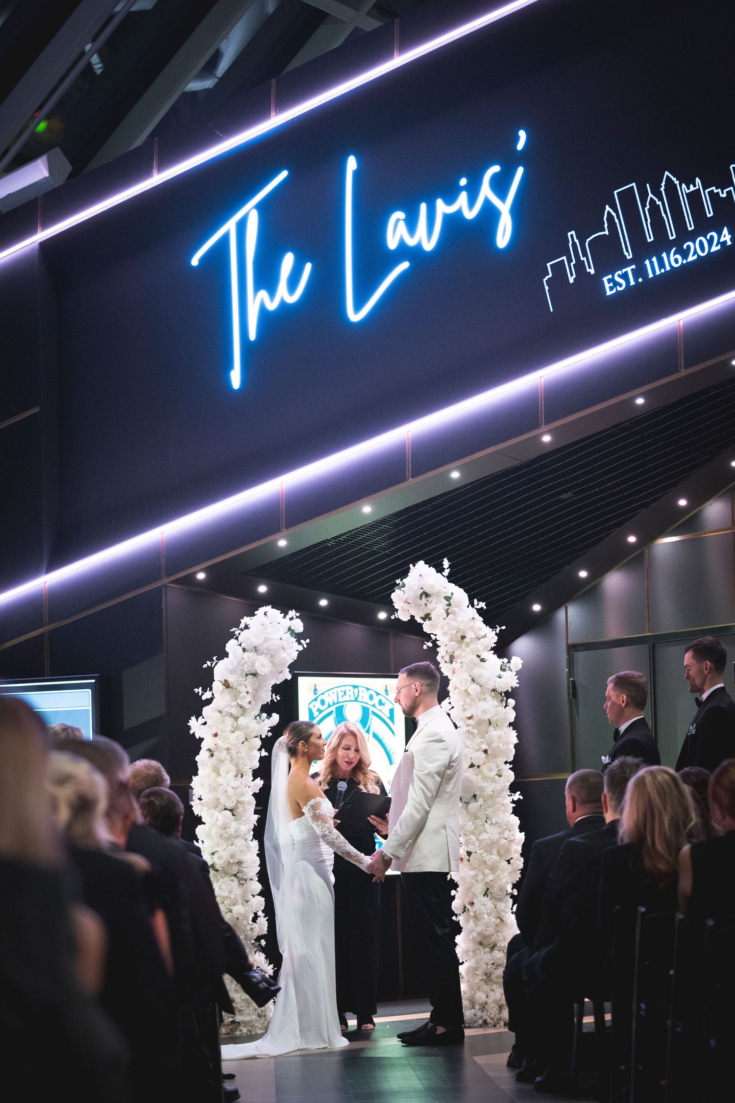 Wedding ceremony with white floral arches and blue neon lighting.