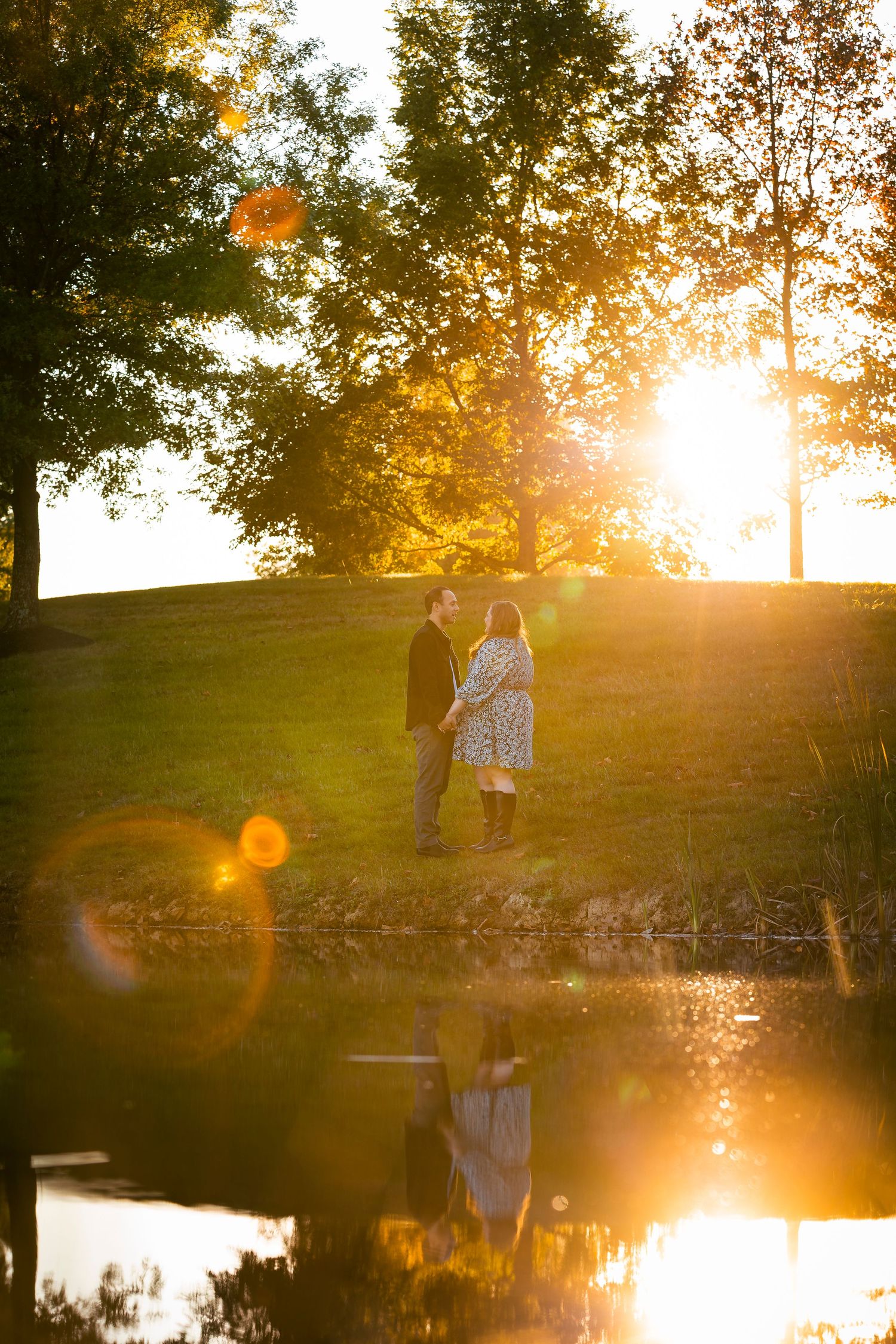 A peaceful pond reflects the golden sunset as two silhouettes stand on its shore in a tranquil natural setting.