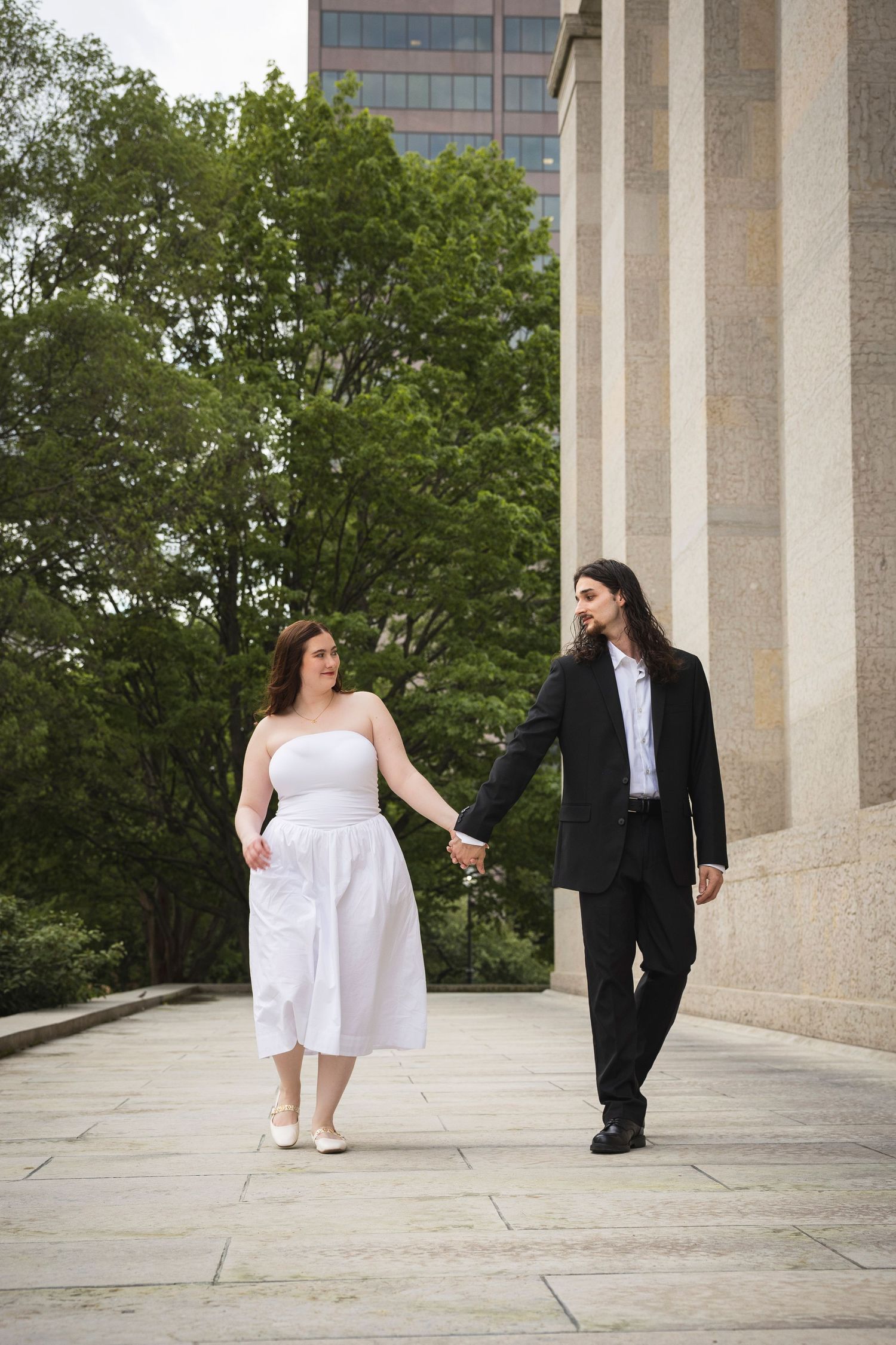 Two people in contrasting black and white attire walk along a colonnaded walkway surrounded by trees.