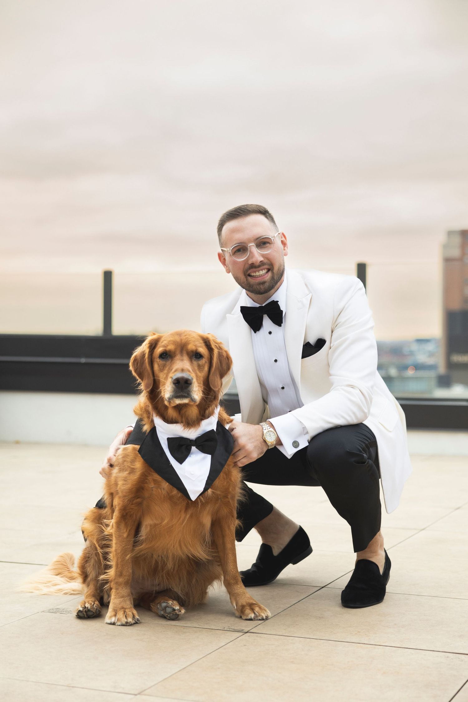 Golden retriever dog wearing a formal bowtie poses with someone in a tuxedo on a rooftop.