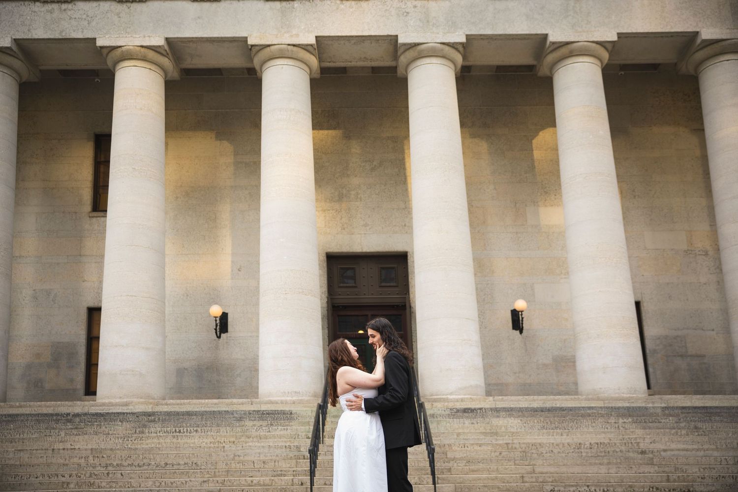 A couple in formal attire shares romantic moments between elegant white columns in a grand architectural setting.