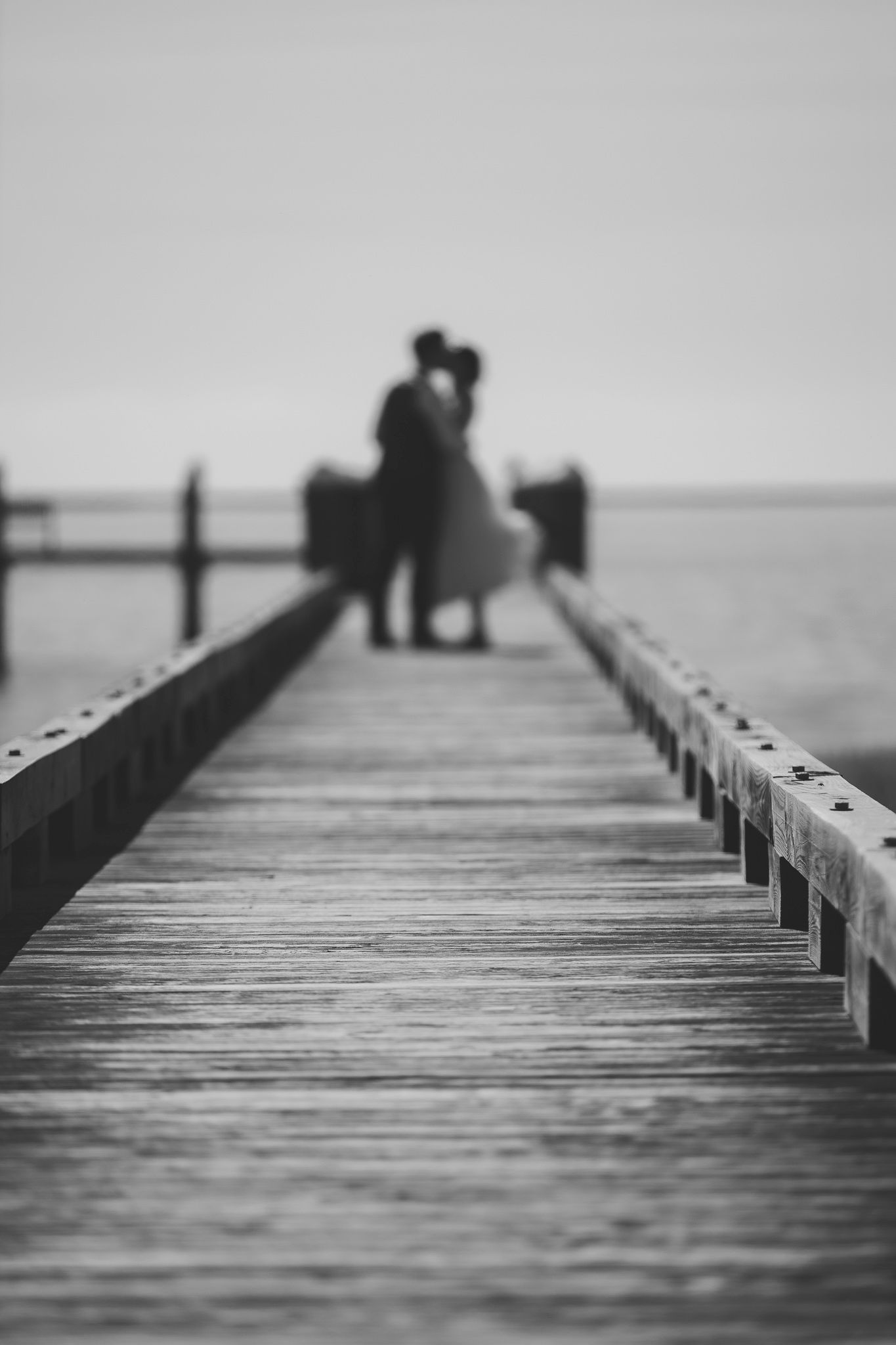 Black and white photo of a wooden pier stretching into misty water with silhouettes in distance.