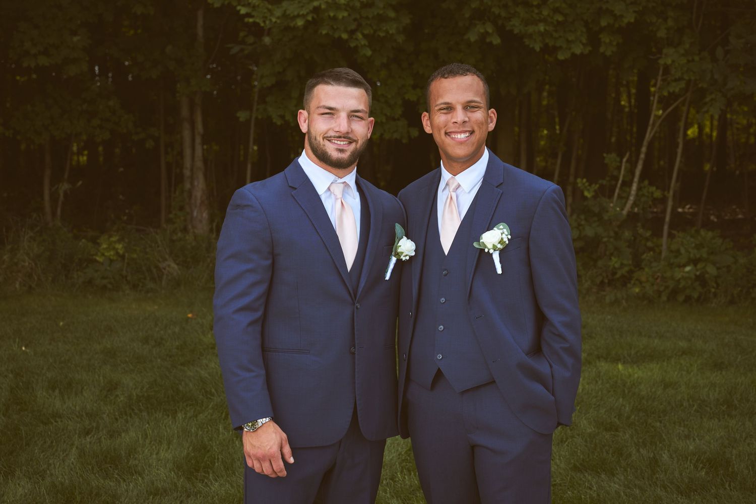 Two groomsmen in navy blue suits with boutonnieres pose together outdoors at a wedding.