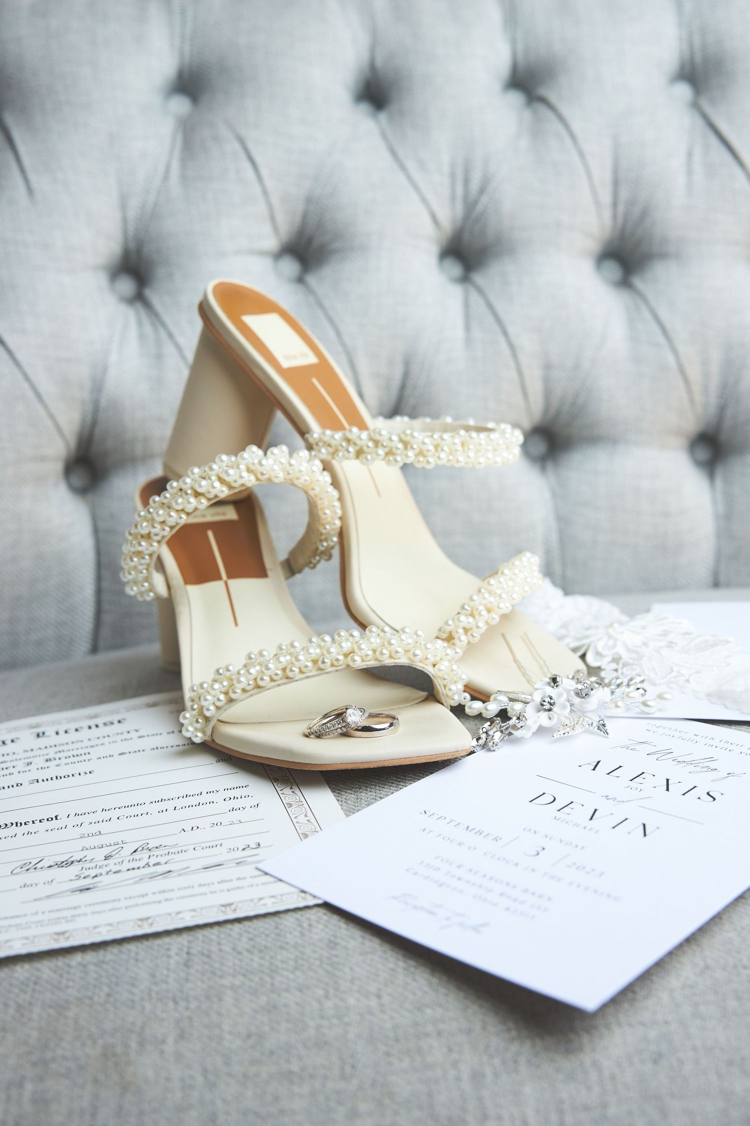 Elegant pearl-embellished bridal shoes displayed with wedding rings and invitation on tufted gray chair.