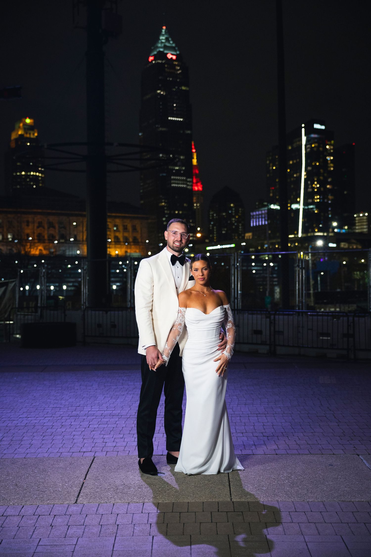 A nighttime wedding photo with a city skyline backdrop and purple-lit pavement.