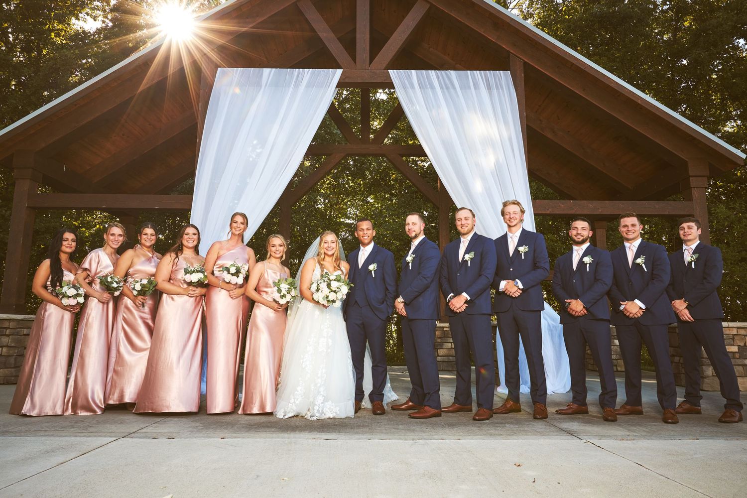 A panoramic view of a wedding party gathered under a wooden pavilion at sunset.