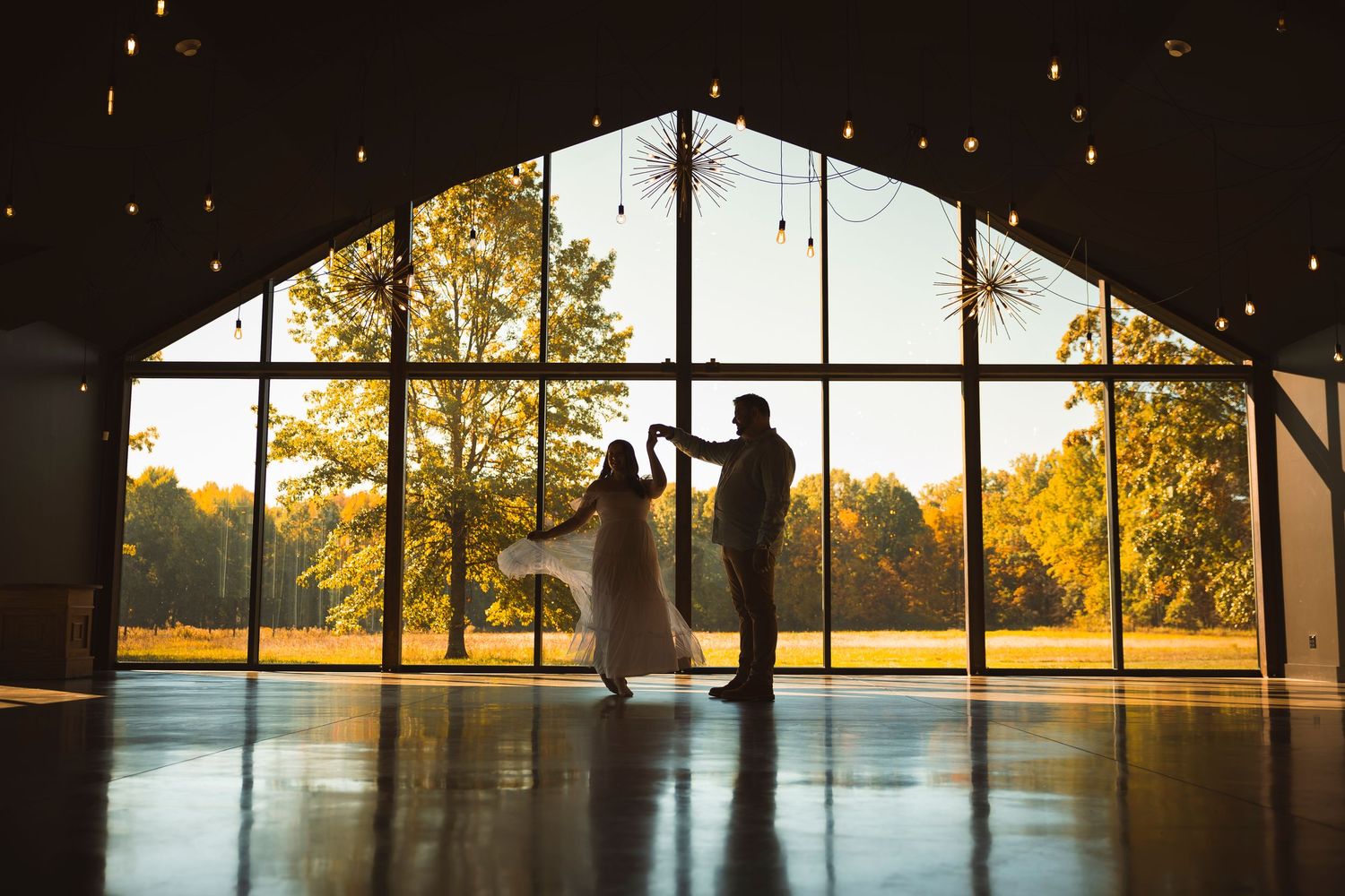 Silhouettes of a couple against large windows with sunset views and string lights.